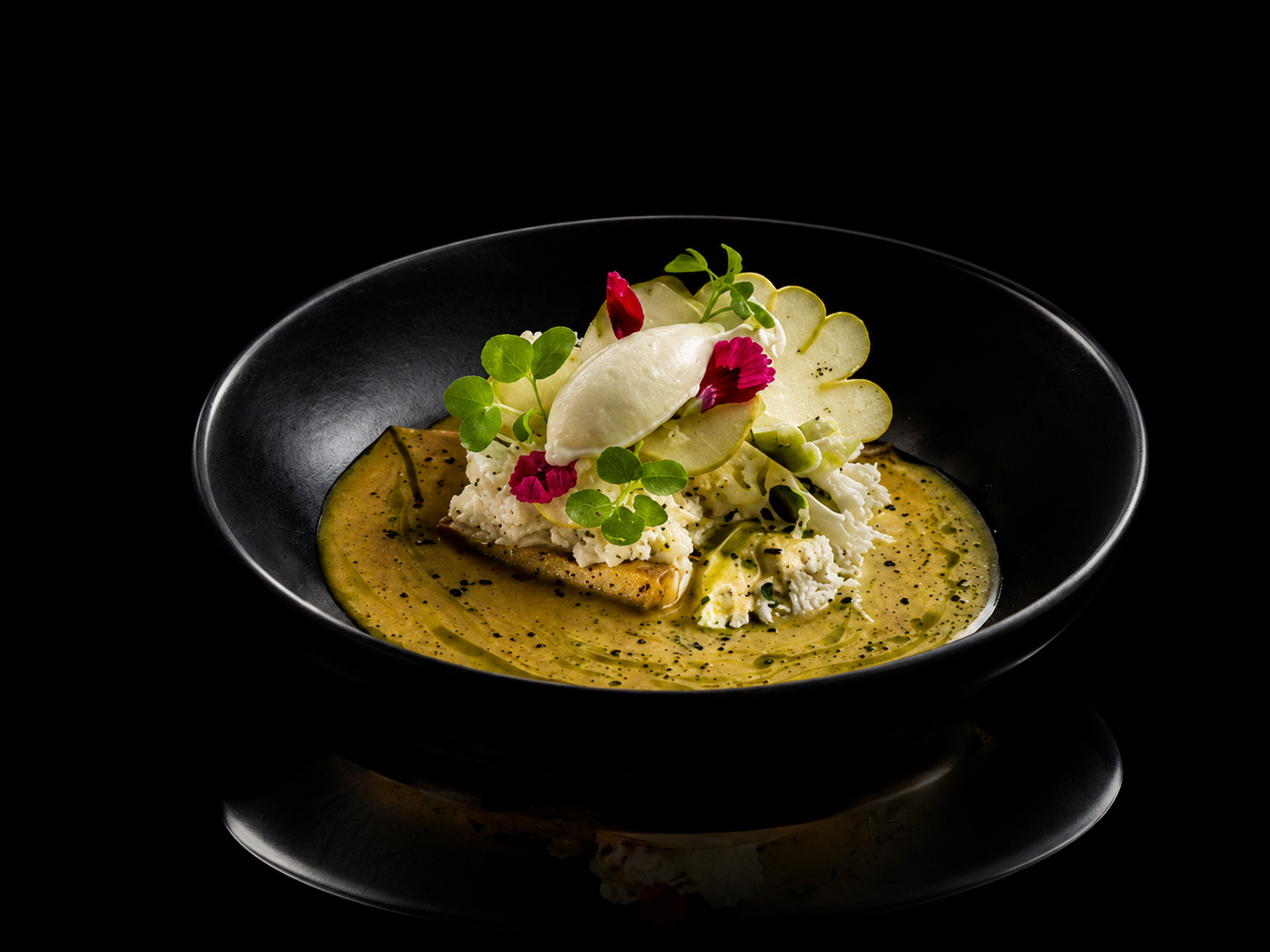 Assiette de cuisine gastronomique avec une purée de céleri, des fleurs comestibles, des rondelles de concombre et une quenelle de crème ou sorbet, présentée dans une assiette noire sur fond noir.