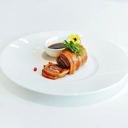 Rôti de viande enroulé, avec une sauce dans un bol, garniture verte et une fleur sur une assiette blanche