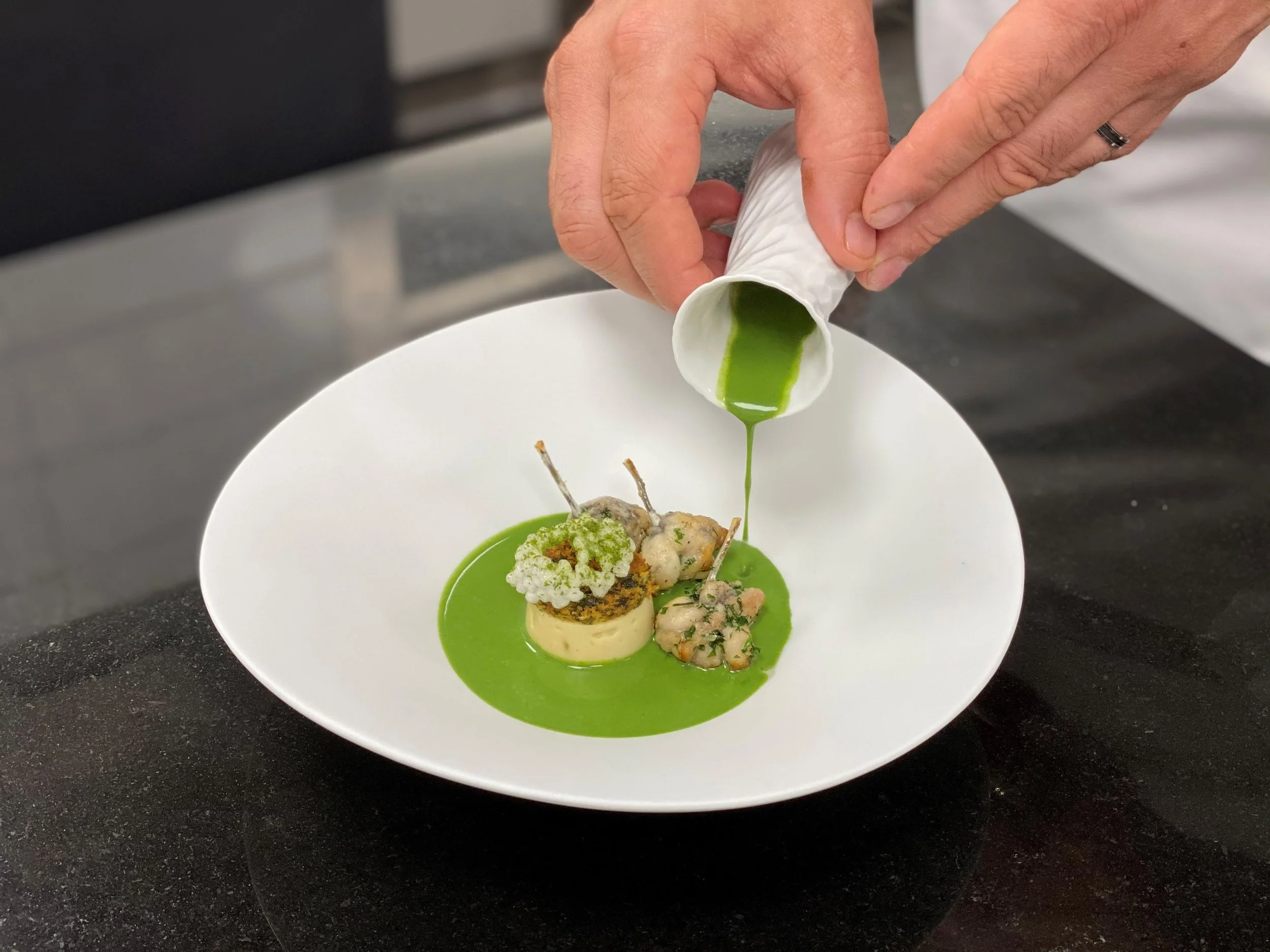 Une personne verse une sauce verte dans une assiette de fine cuisine, contenant plusieurs petits plats et garnitures.