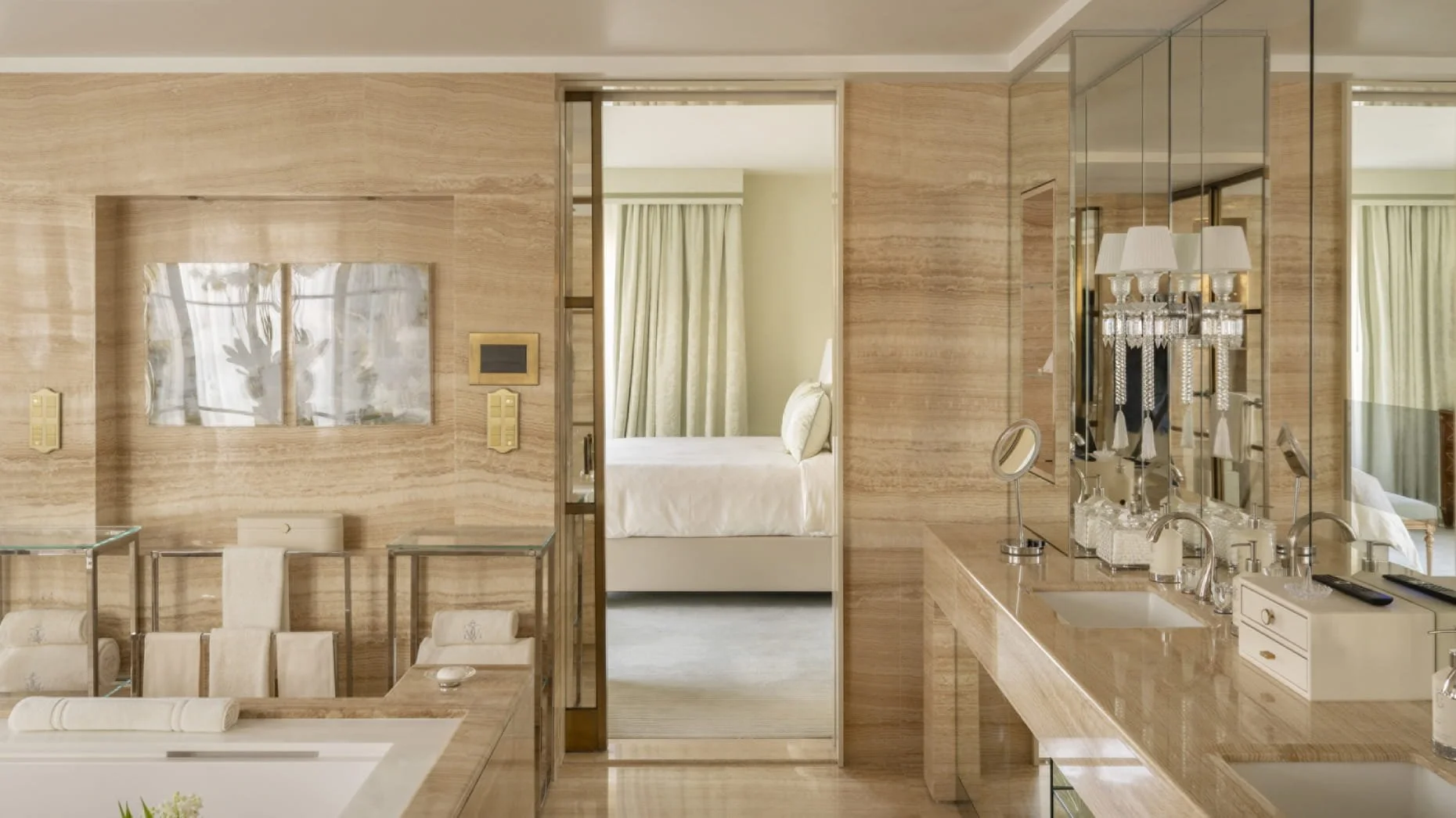 Intérieur d'une chambre d'hôtel élégante avec un mur en marbre beige, un lit blanc visible à travers une porte ouverte, et un coin salle de bain avec miroir, lavabo et lampes à cristaux.