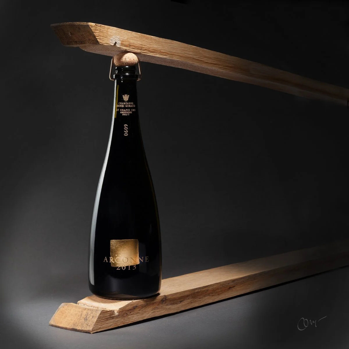 Une bouteille de champagne Arcoine 2013 équilibrée sur une poutre en bois, avec une autre poutre en bois reposant en angle entre la bouteille et le fond noir.