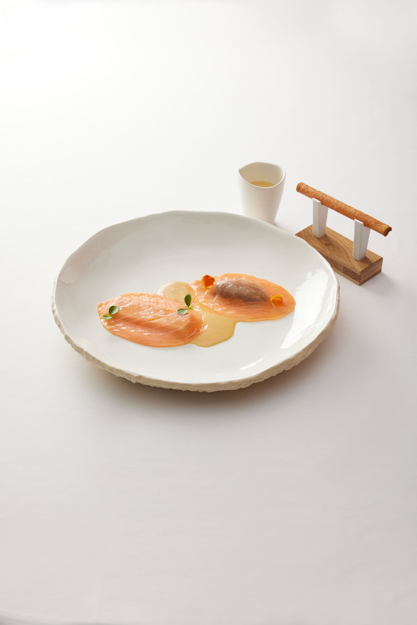 Assiette avec deux filets de saumon cru, garnitures et sauce dans un cadre minimaliste, avec un petit verre et un bâtonnet de pâte à côté.