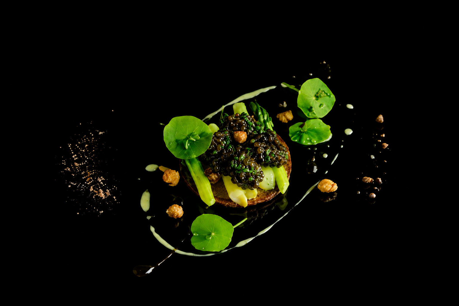 Présentation d'un plat gastronomique avec des éléments verts, des couverts de caviar, et une sauce blanche sur fond noir.