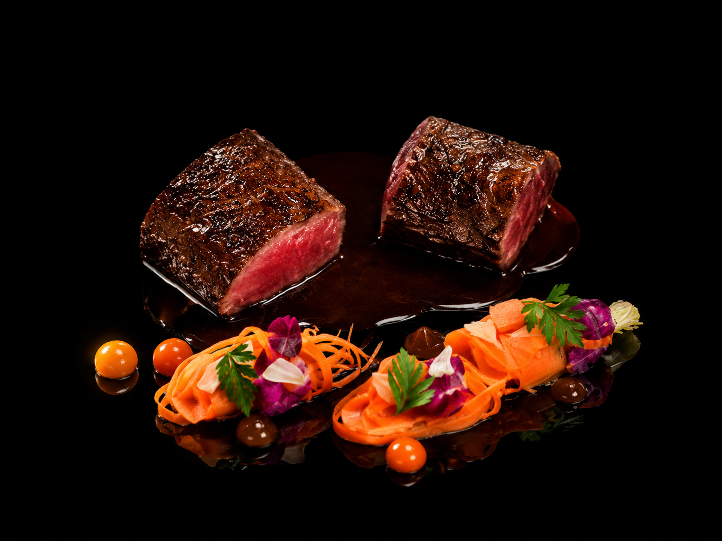 Deux morceaux de steak mi-saignant avec sauce, accompagnés de carottes râpées, fleurs comestibles, et petites sauces colorées, présentés élégamment sur une surface noire.