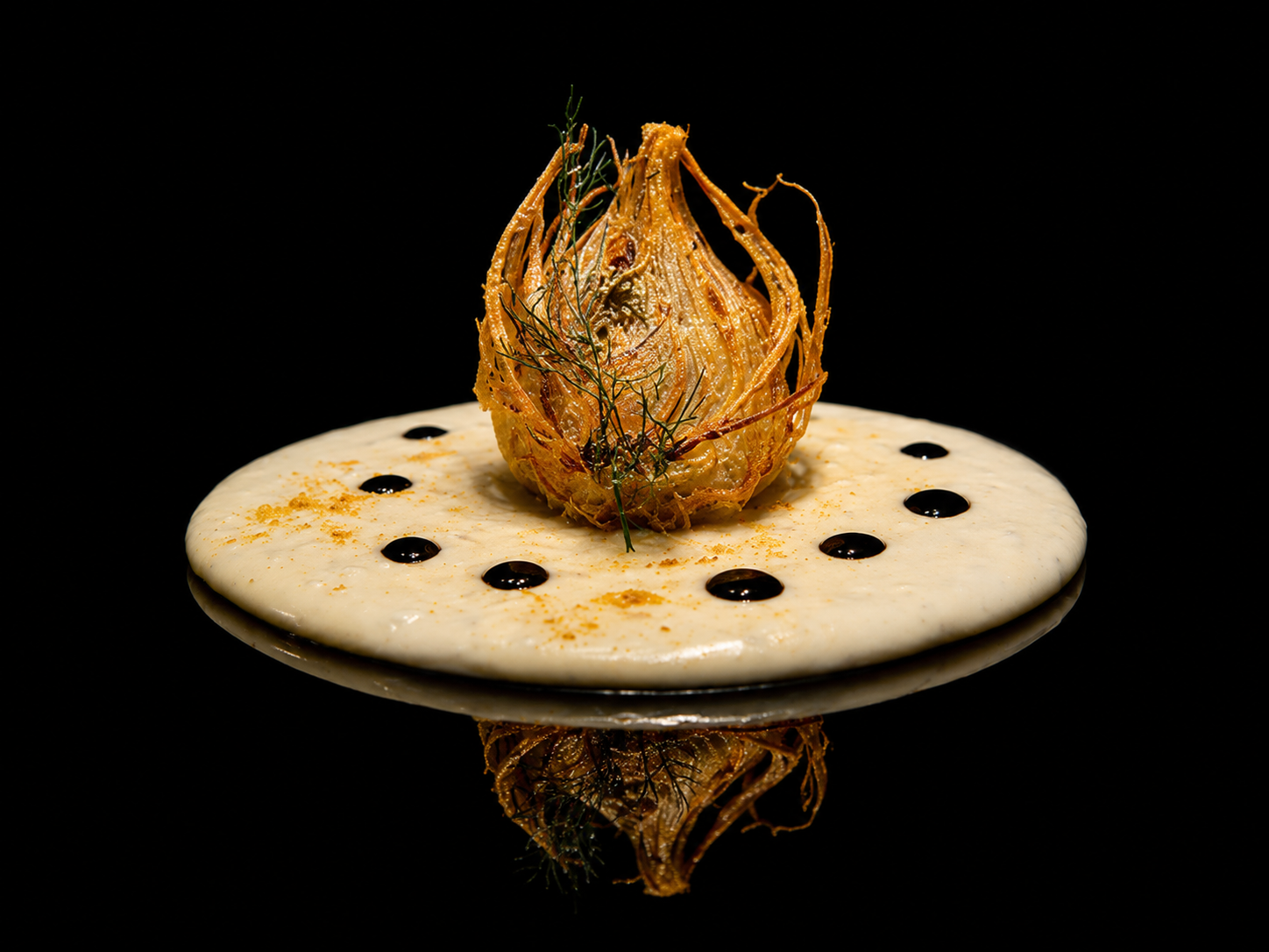 Plat gastronomique avec un croustillant en forme de fleur, servi sur une sauce blanche avec des gouttes de sauce sombre, présenté sur une surface noire.