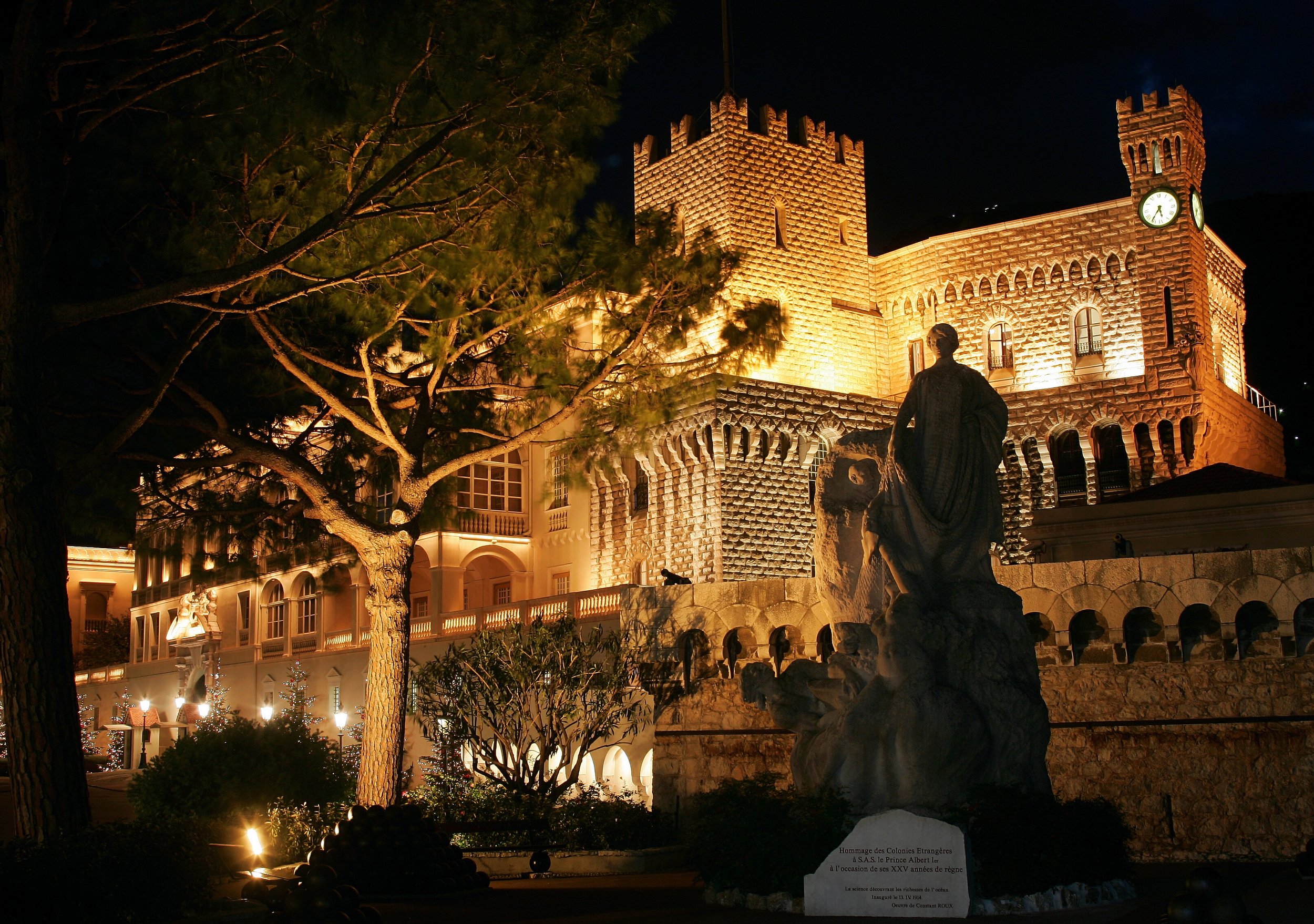 Château illuminé la nuit avec statue en premier plan, arbres et lampadaires dans le jardin.