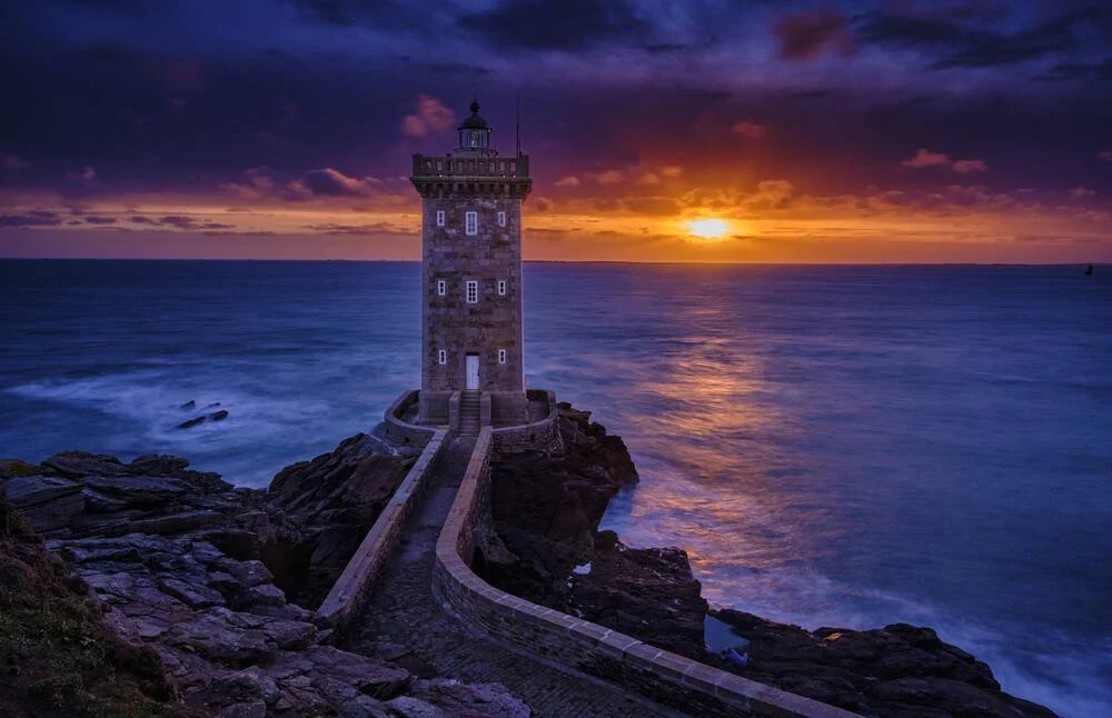 Phare en pierre sur une côte rocheuse au coucher du soleil, avec un ciel orageux et la mer agitée.