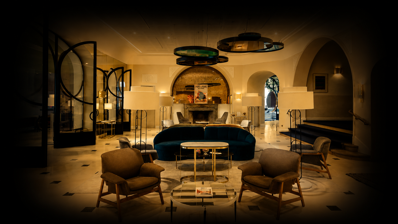 Salon intérieur élégamment décoré avec des fauteuils, une cheminée en pierre, des lampes sur pied, et une scène centrale avec un sofa bleu foncé, une table basse, et des œuvres d'art au mur, éclairé chaleureusement.