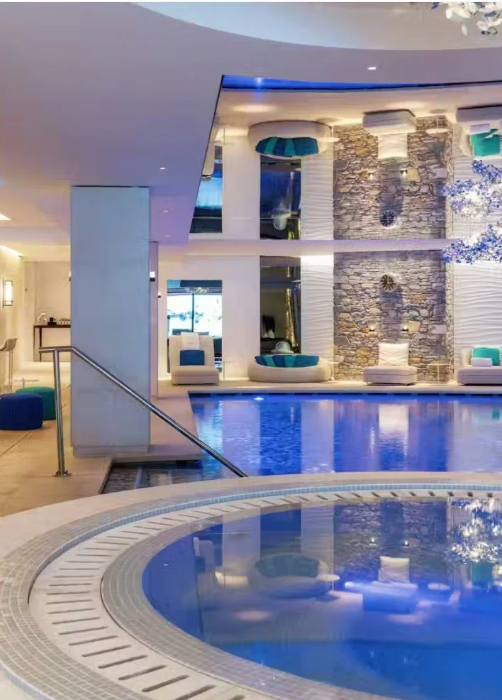 Piscine intérieure avec des sièges confortables, un mur en pierre décoratif, et un plafond moderne avec éclairage indirect dans une ambiance relaxante.