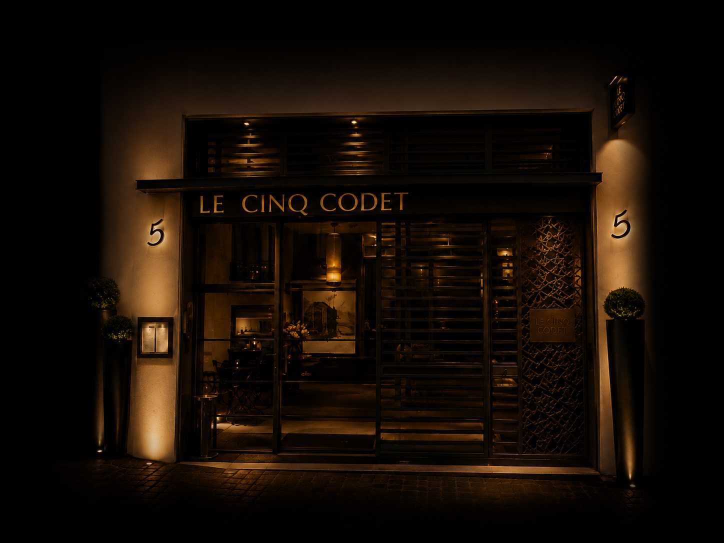 Façade d'un restaurant appelé 'Le Cinq Codet' avec un éclairage tamisé nocturne, deux plantes en pot de chaque côté de l'entrée, et une porte en verre avec le nom du restaurant affiché.