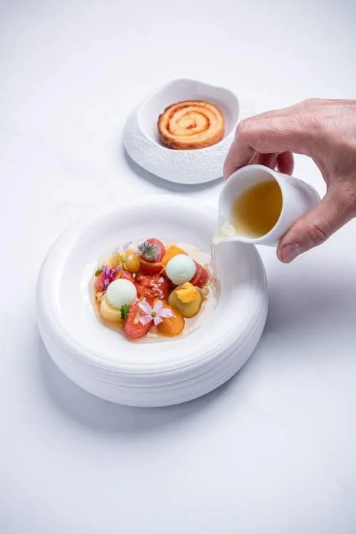 Une main verse de la crème ou du liquide sur une assiette de dessert décoré de fruits, d'œufs de poisson, et de fleurs, avec un biscuit enroulé sur une petite assiette à côté.