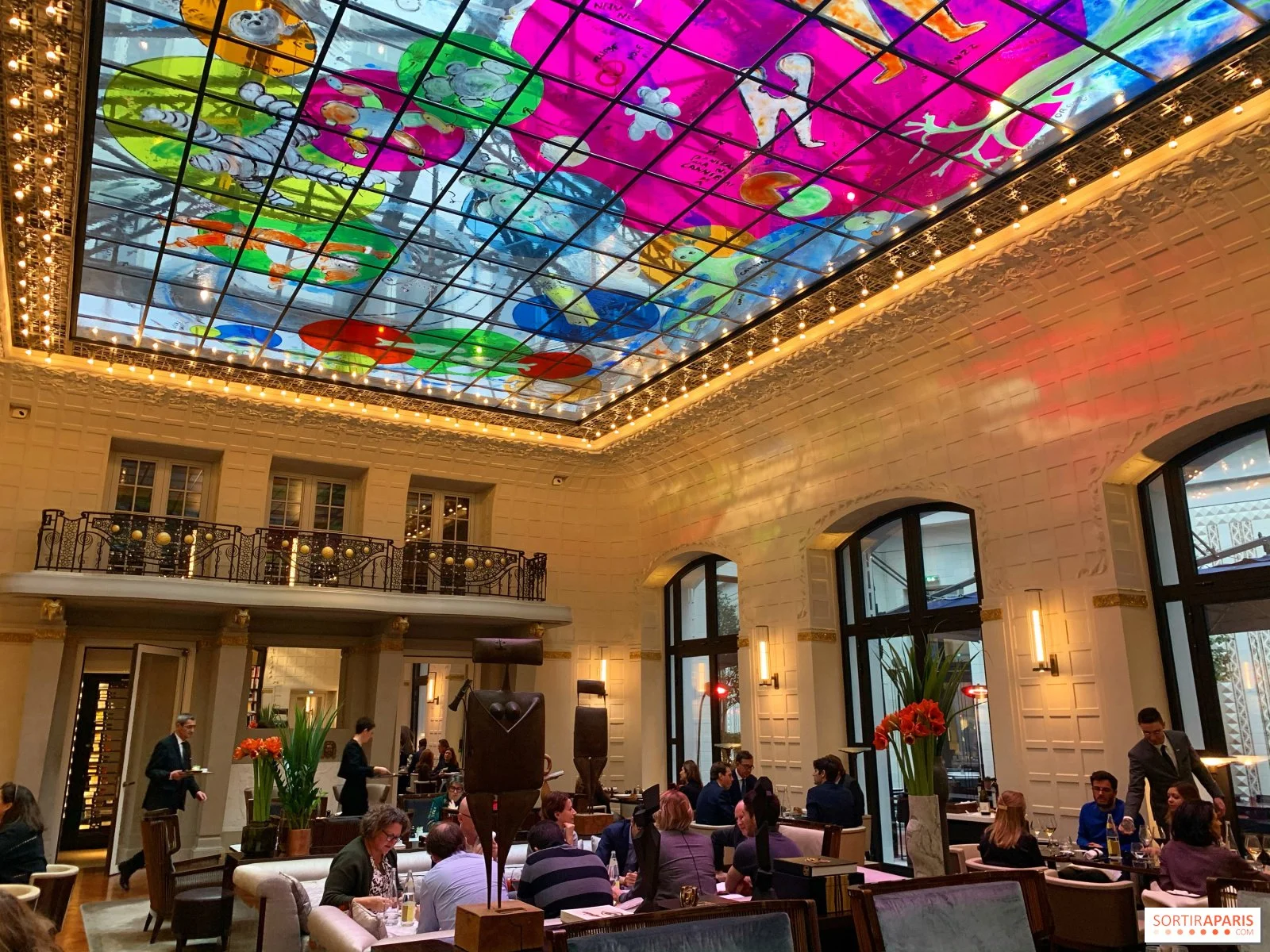 Une salle de restaurant élégante avec un plafond en verre coloré et une ambiance chaleureuse. On y voit des tables avec des clients et des serveurs en service, des fleurs rouges en décoration, et des fenêtres arrières en arqué.