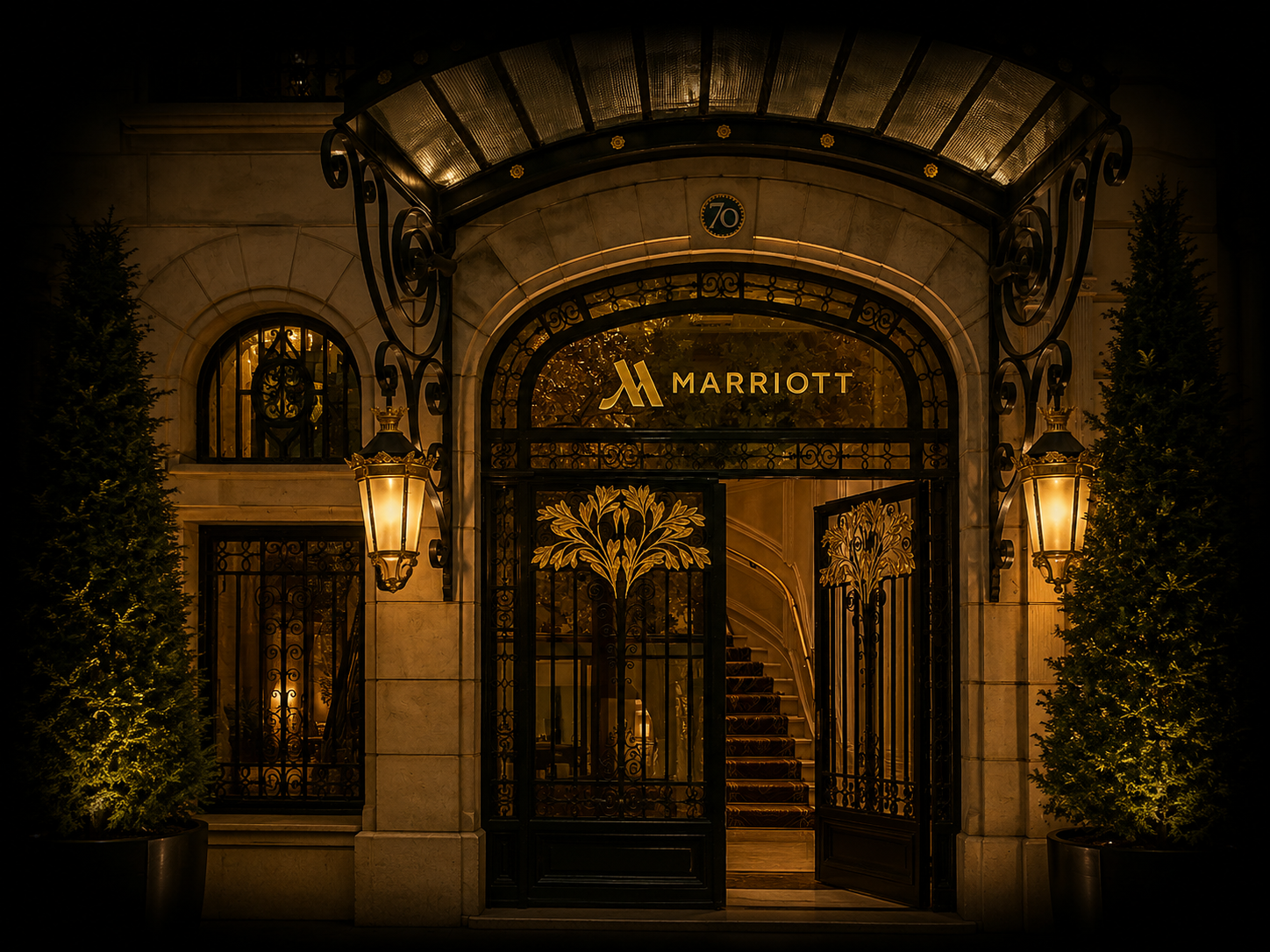 Entrée élégante de l'hôtel Marriott, éclairée par deux lampadaires et entourée de deux arbres de Noël, avec un portail en fer forgé et un escalier intérieur visible par la porte.