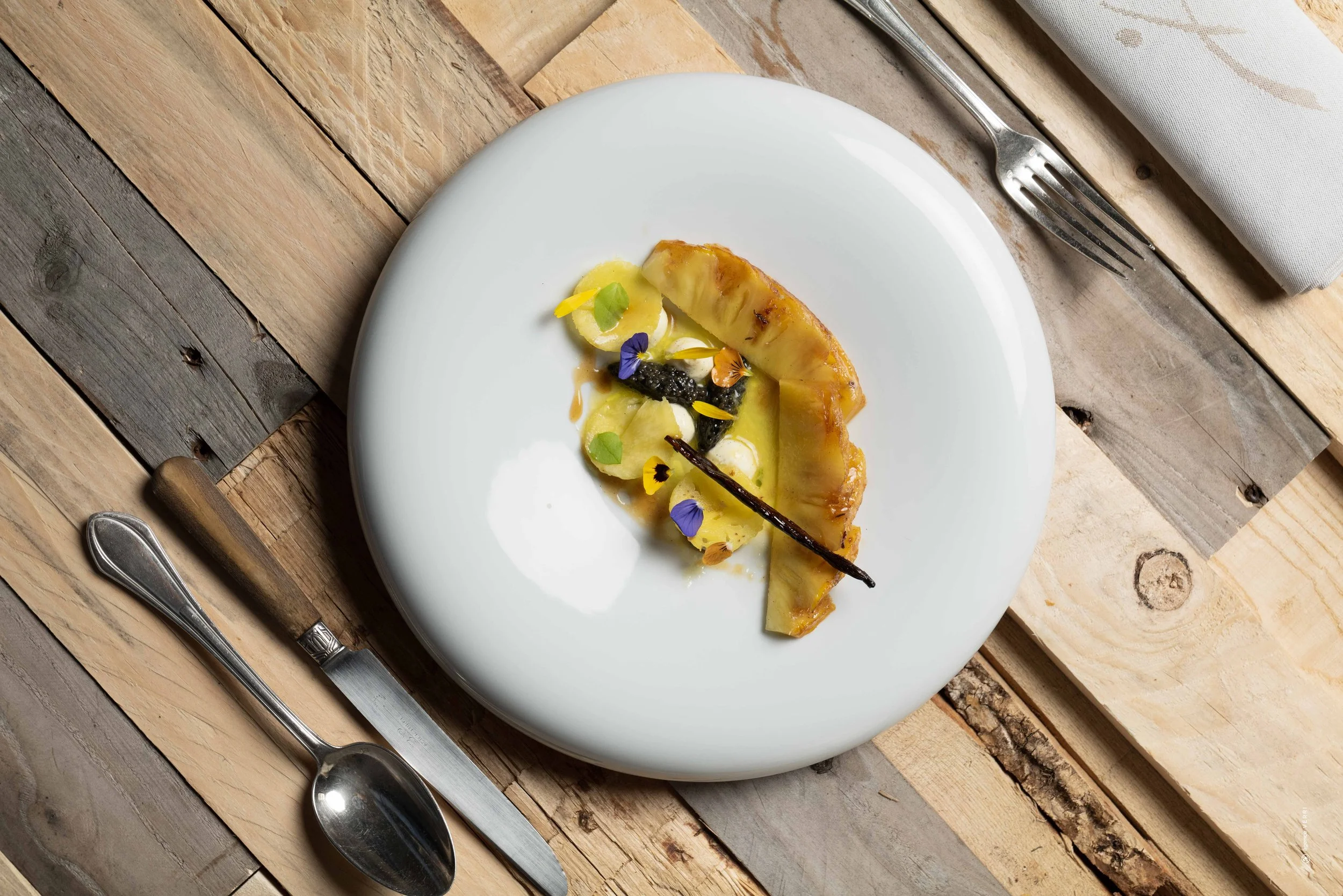 Assiette de dessert avec une tranche de ananas caramélisé, accompagnée de fruits, fleurs edulcorés et une sauce jaune, sur une table en bois avec fourchette, couteau et nappe blanche.