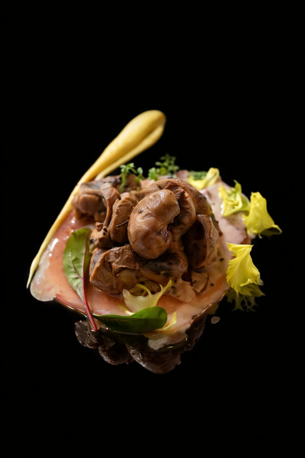 Plat de viande avec champignons, servi avec une sauce, décoré de feuilles et fleurs comestibles, sur un fond noir.