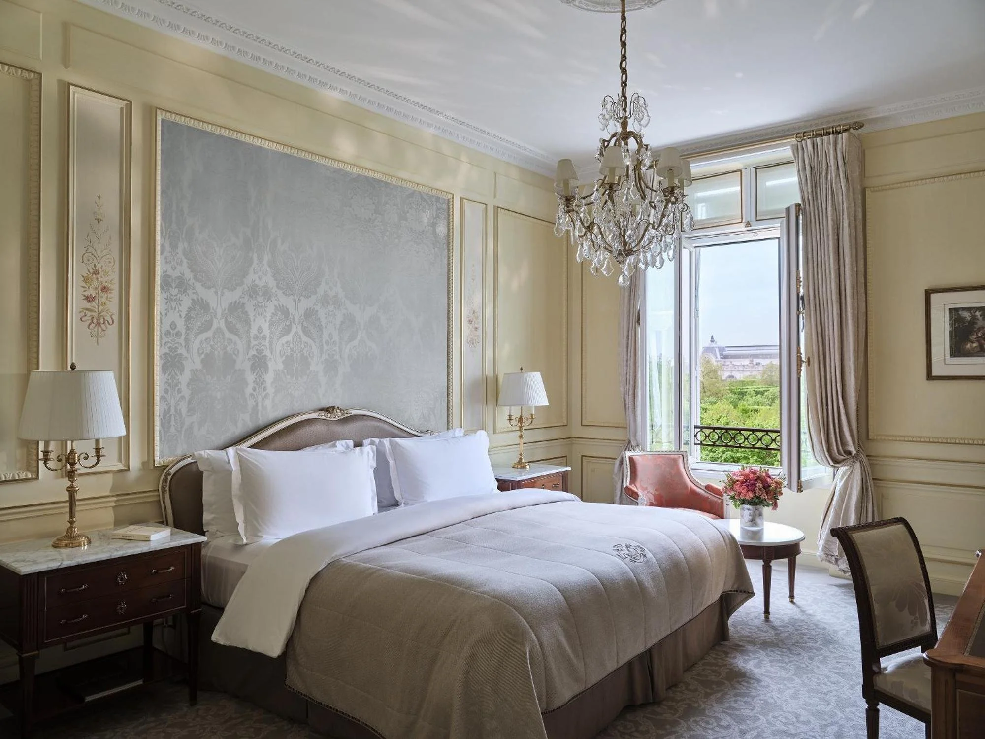 Chambre élégante avec un grand lit, lampes de chevet, chaises, table avec un bouquet de fleurs, grande fenêtre laissant entrer la lumière, rideaux en tissus clair, décoration classique et chandelier en cristal.