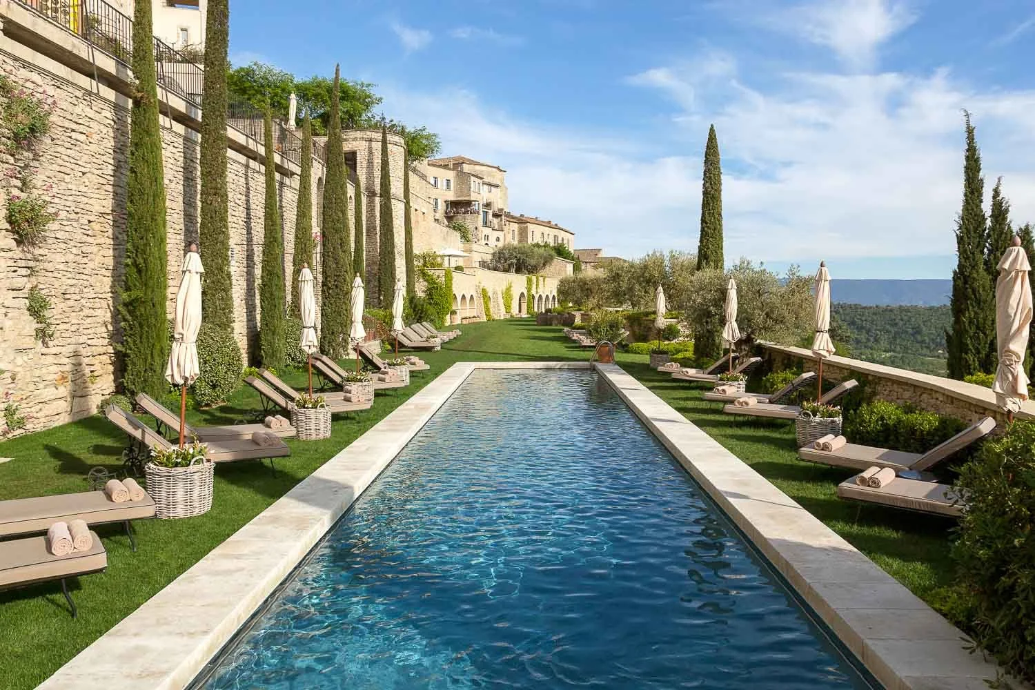 Vue d'une piscine à débordement entourée de chaises longues avec serviettes, sous un ciel bleu, avec des cactus et des arbres en arrière-plan dans un paysage vallonné.