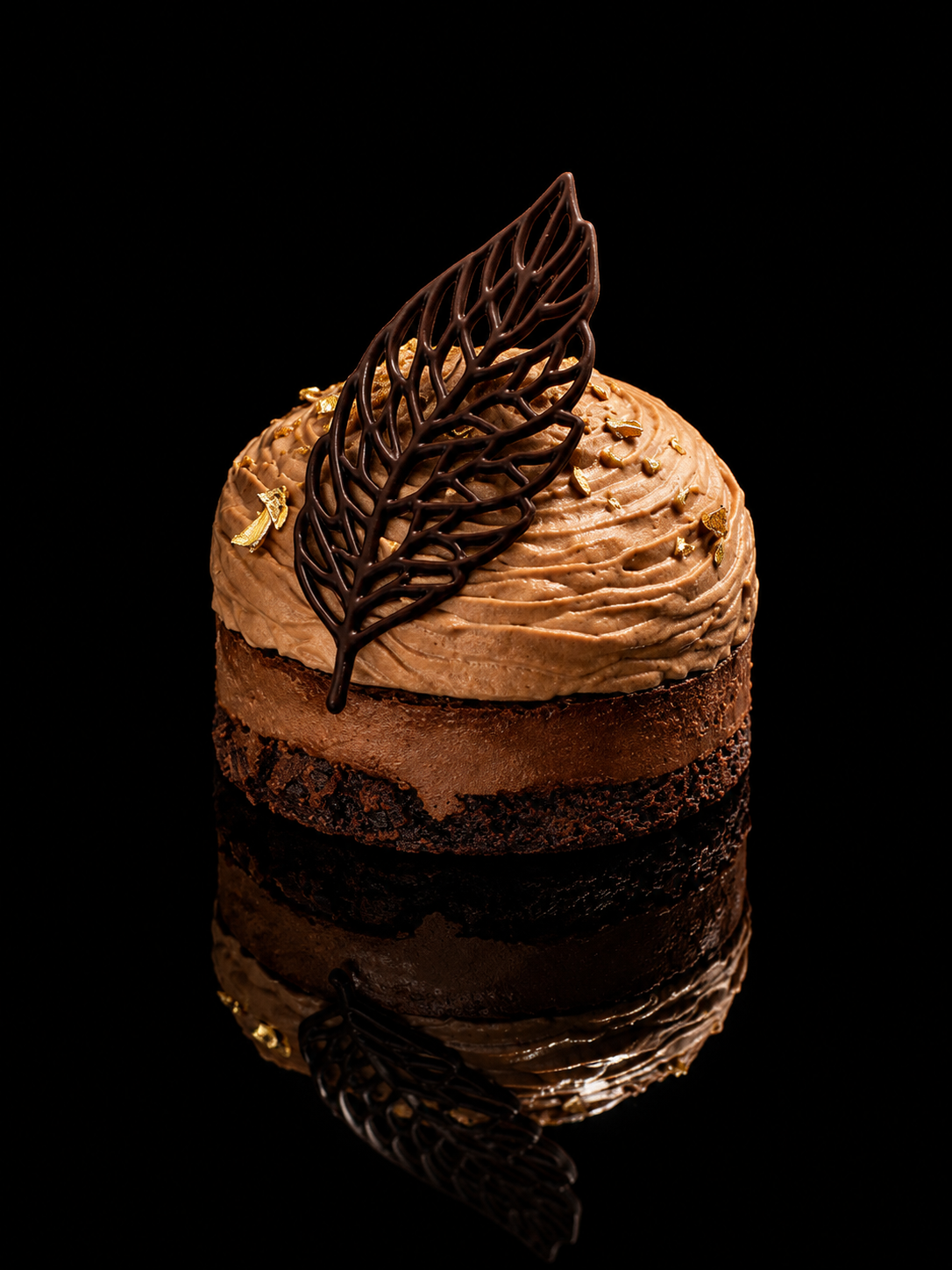 Gâteau au chocolat avec décoration en chocolat, une feuille en chocolat et des éclats dorés, sur fond noir reflété en dessous.