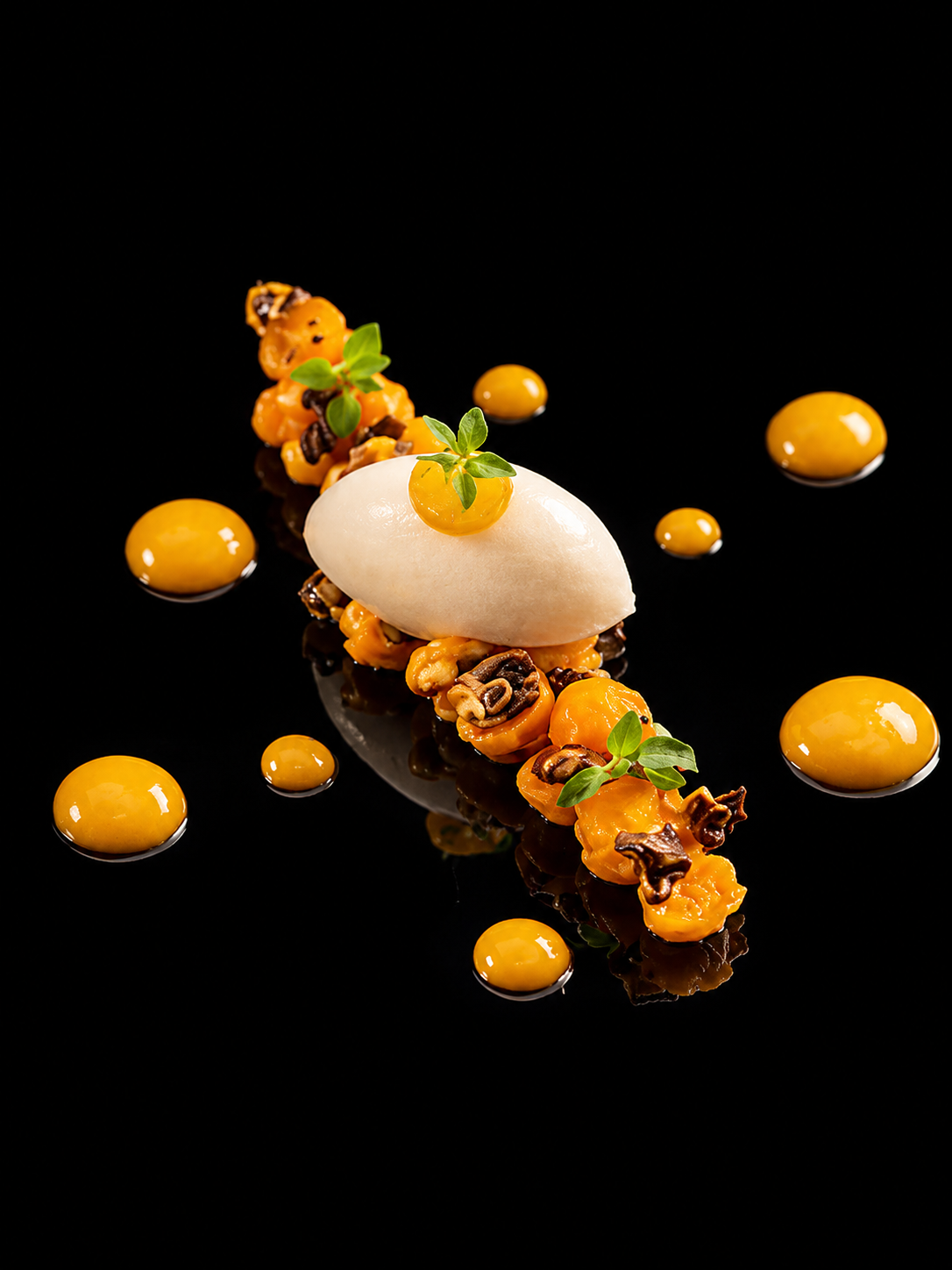 Assiette de dessert moderniste avec une quenelle de glace blanche, des morceaux d'orange, des micro-pousses, et une sauce caramel, présentée sur un fond noir.