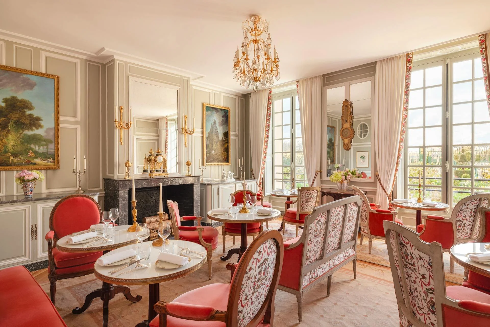 Salon élégant avec murs en bois clair, tableaux, cheminée en marbre, chandeliers et grandes fenêtres avec rideaux roses et blancs, mobilier en bois et tissu rose et floral.