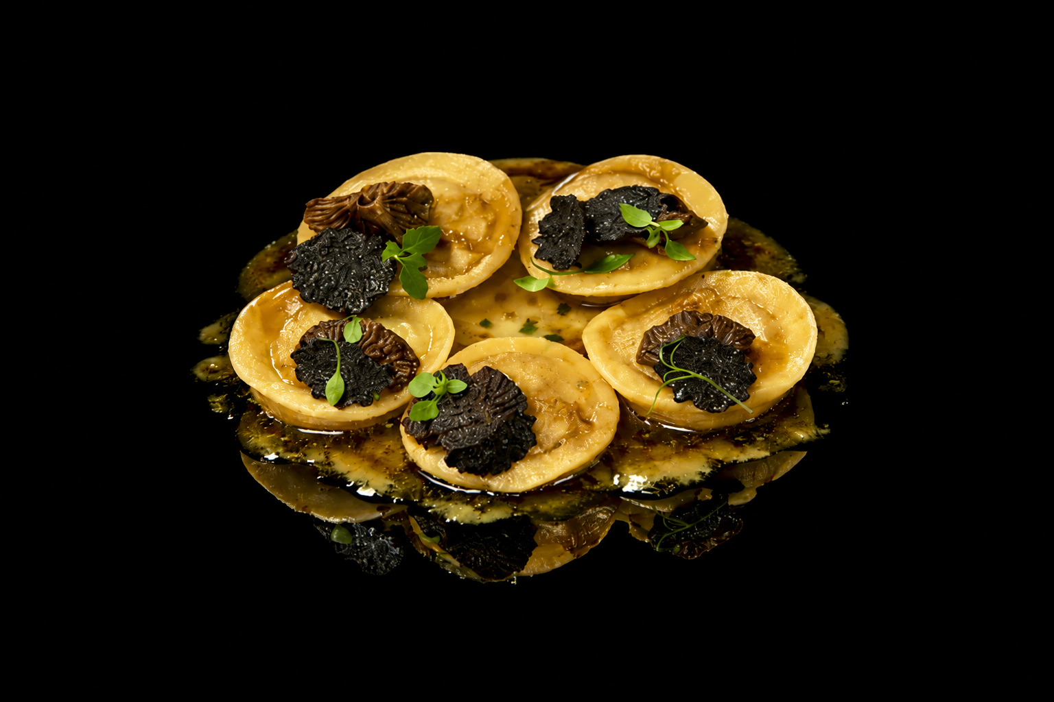 Assiette de raviolis garnis de truffes noires et de microgreens sur une surface noire réfléchissante.