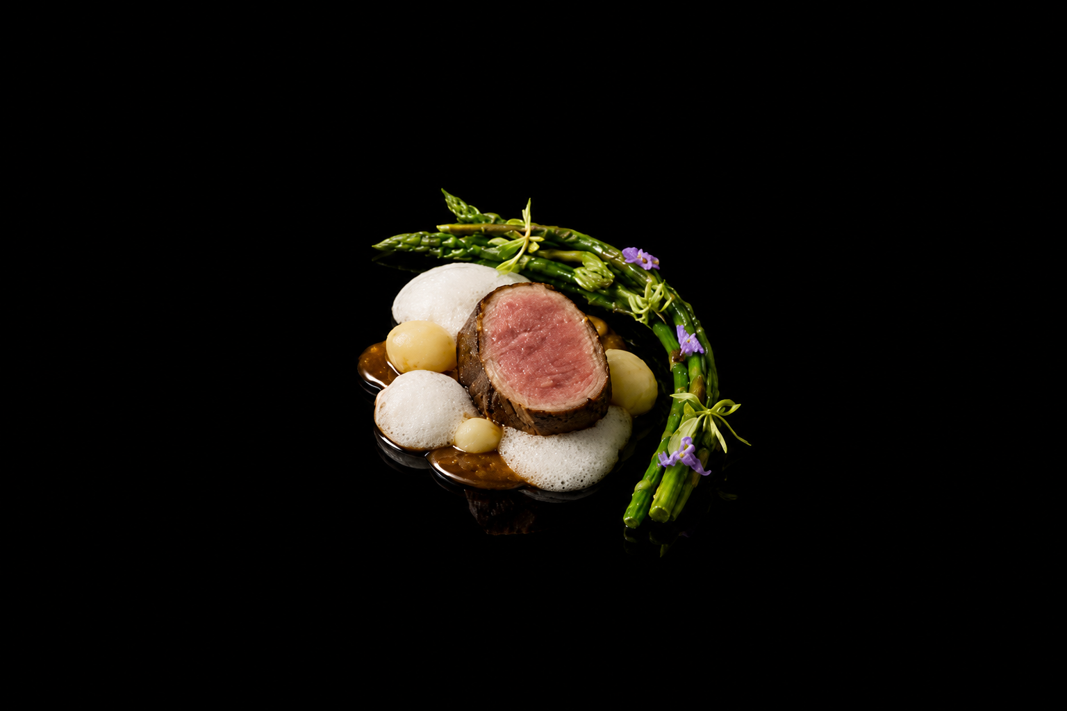 Assiette gastronomique avec un filet de viande cuite à point, accompagnée de asperges, petites pommes de terre, mousse blanche, sauce, et décorations florales, sur fond noir.