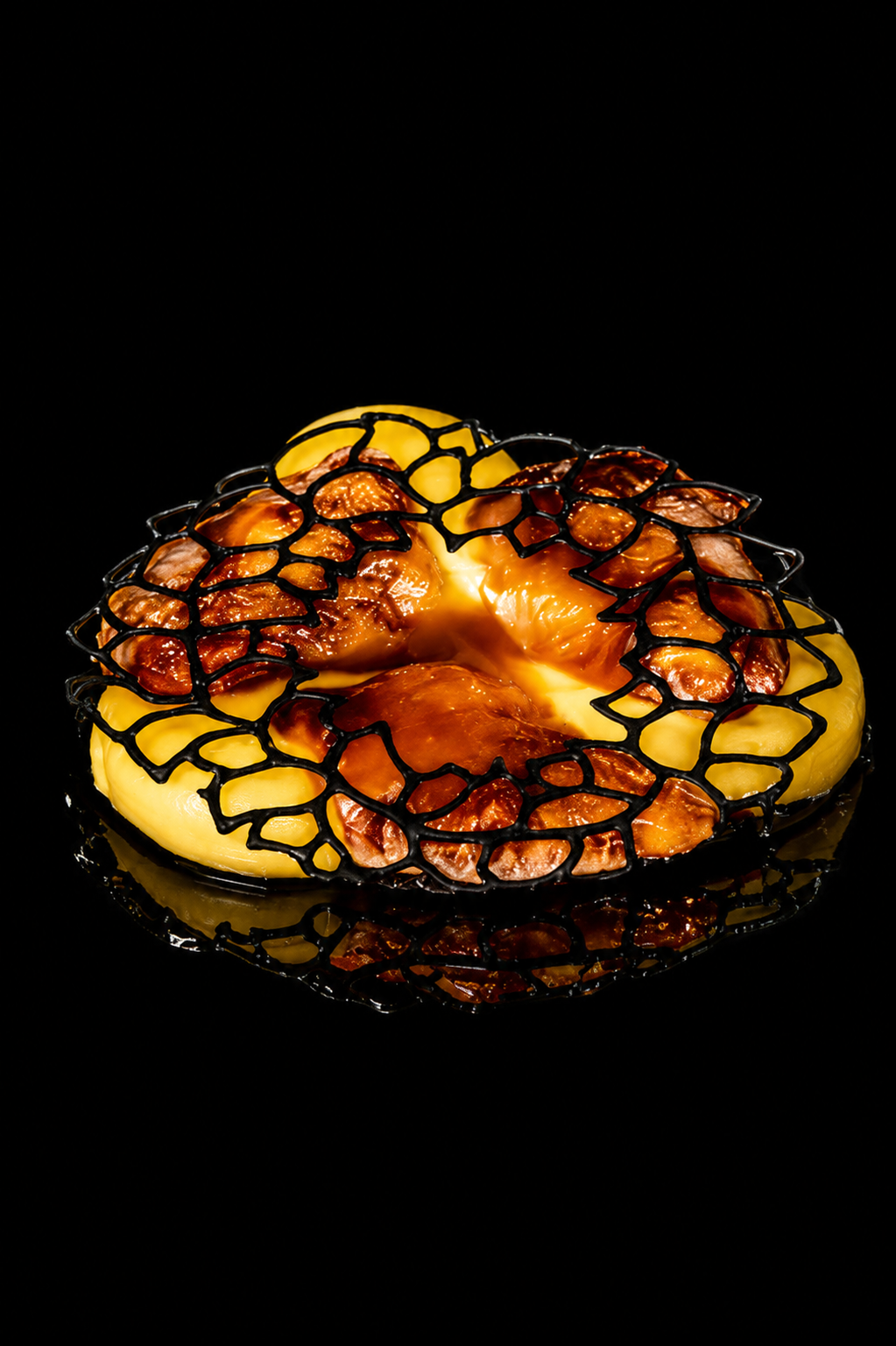 Un dessert avec un donut jaune recouvert de caramel et de morceaux de fruits, décoré avec un filet noir en forme géométrique, reflet du dessert sur une surface noire.