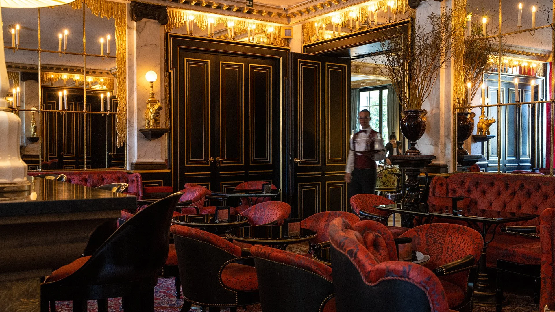 Intérieur d'un restaurant ou salon avec des meubles en velours rouge, moulures dorées, miroirs, lampes à bougies, et un serveur portant un plateau.