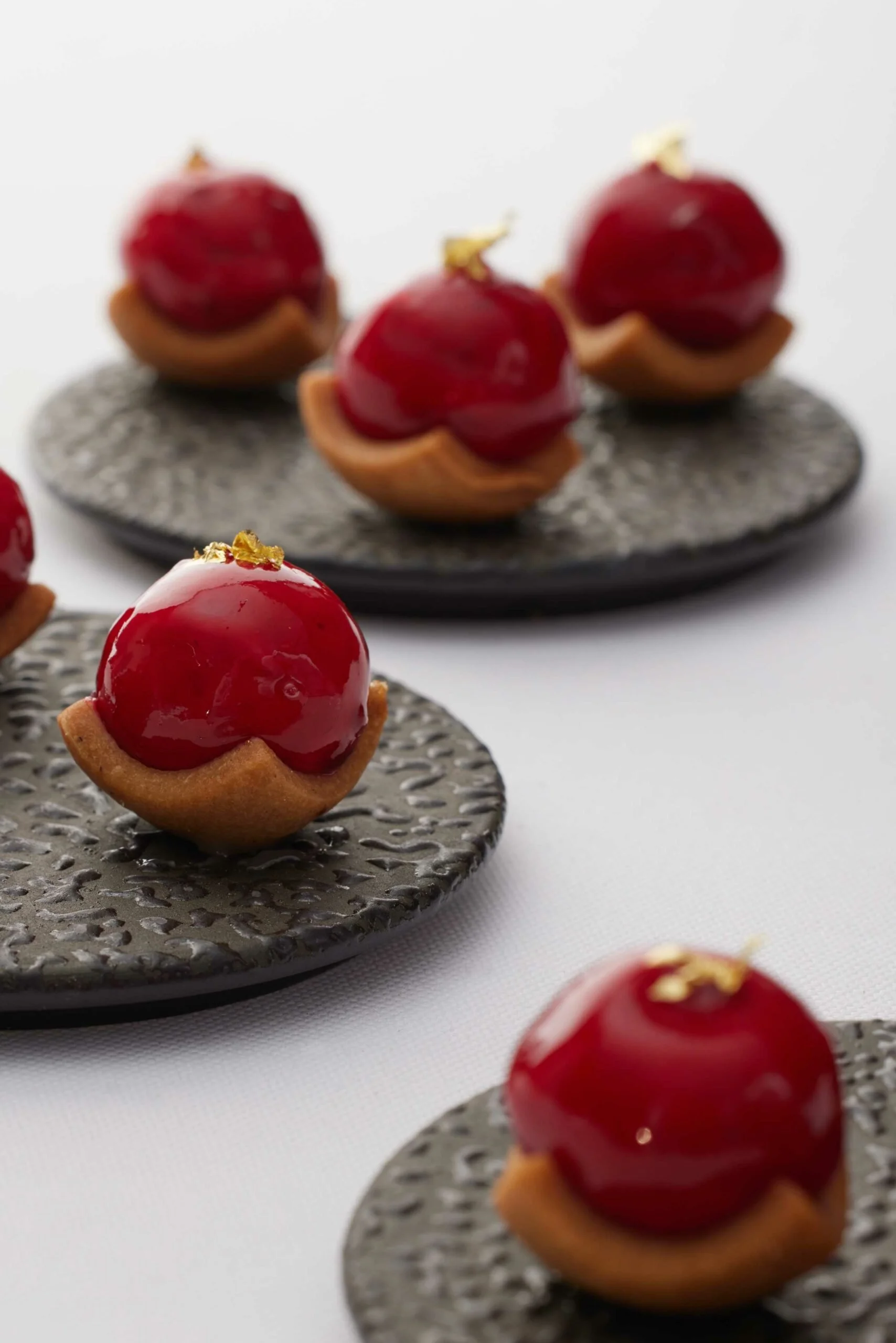 Petites tartes avec une base en biscuit, garnies d'une crème et d'une cerise brillante, décorées de feuilles d'or, présentées sur des assiettes en pierre