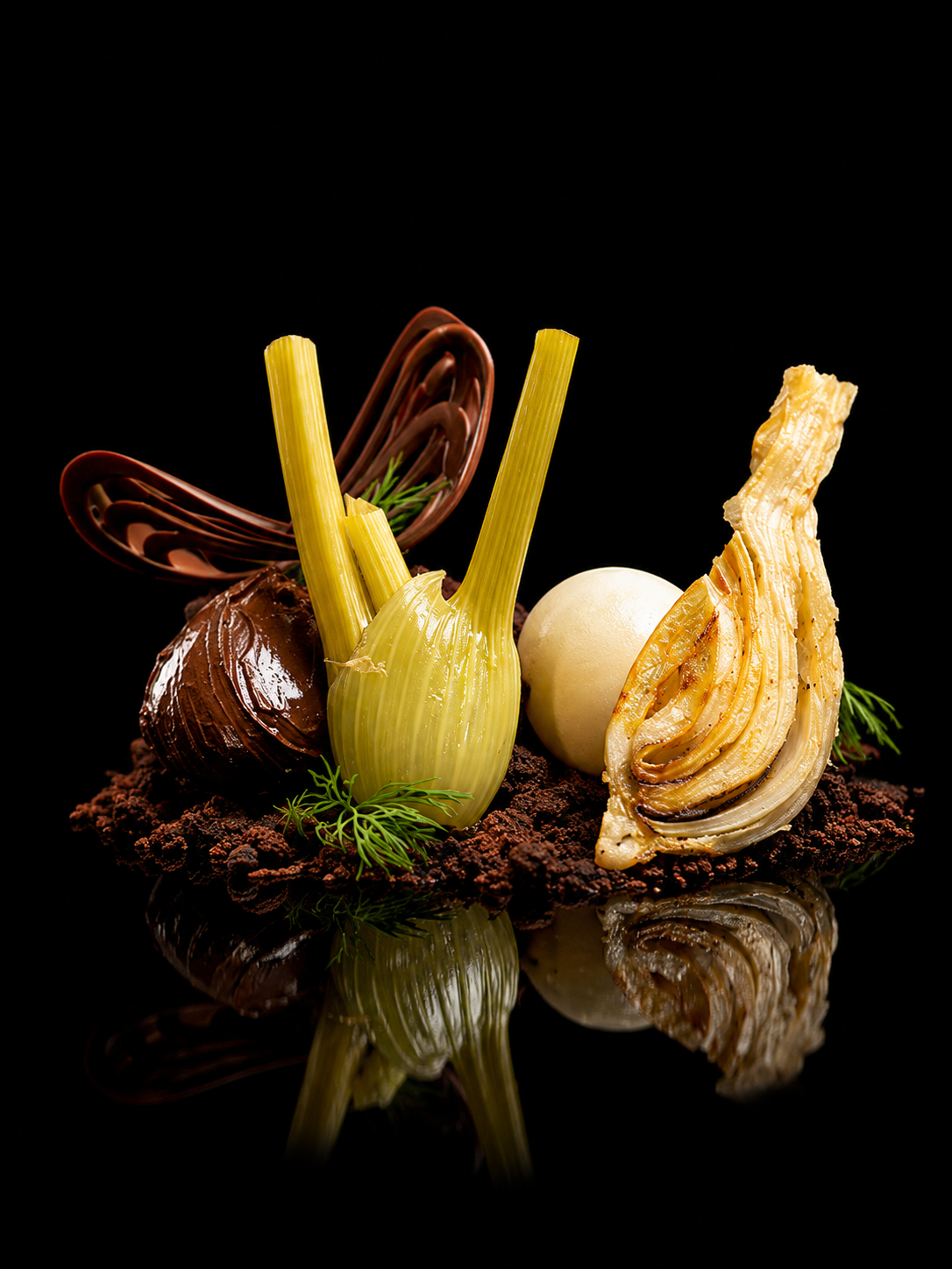 Gâteau ou dessert sophistiqué avec des éléments décoratifs en chocolat, un œuf blanc, et des légumes comme de l'ail. Charme visuel avec des reflets sur une surface noire.