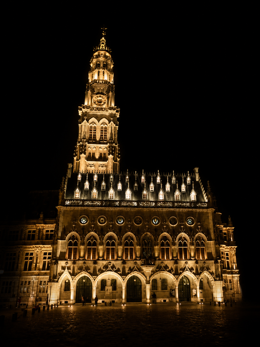 Une grande cathédrale gothique illuminée la nuit, avec un clocher haut et une façade ornée de plusieurs fenêtres en arche et de détails architecturaux complexes.