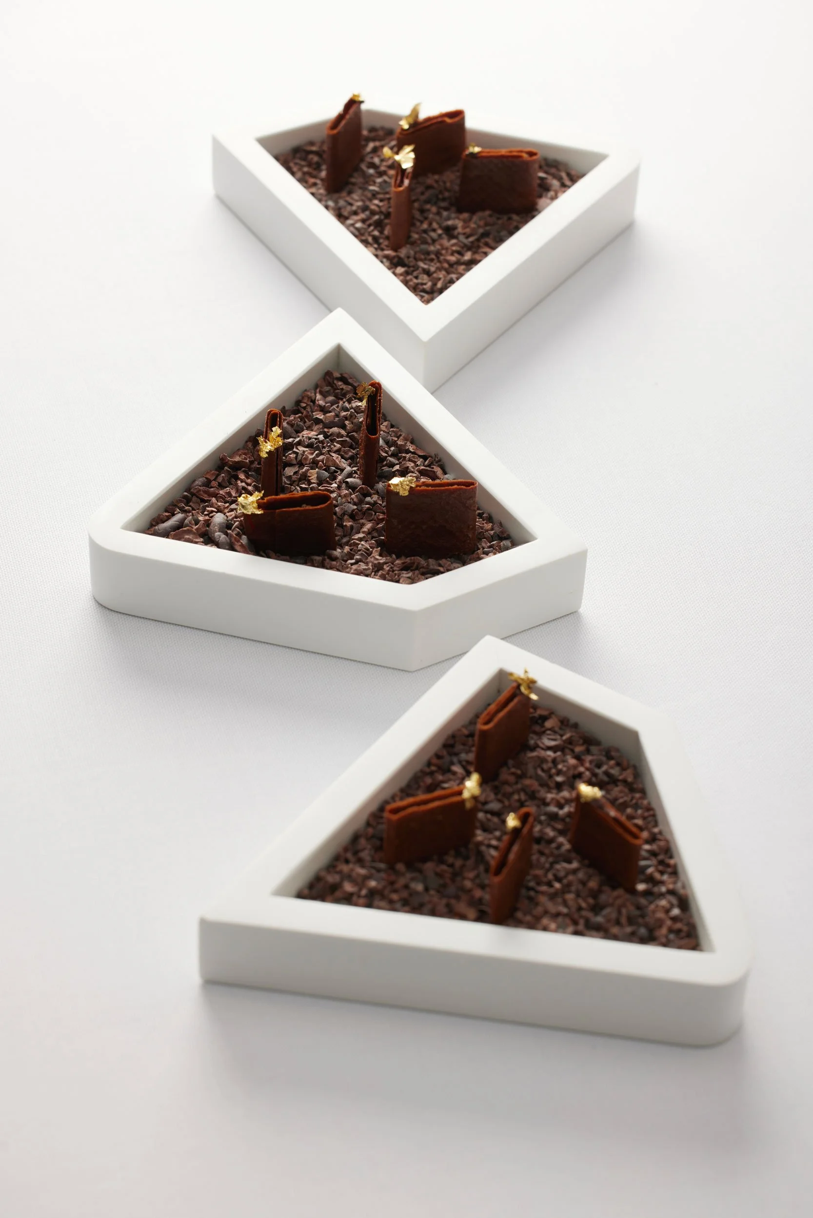 Trois plats triangulaires blancs avec des morceaux de chocolat et des feuilles d'or.