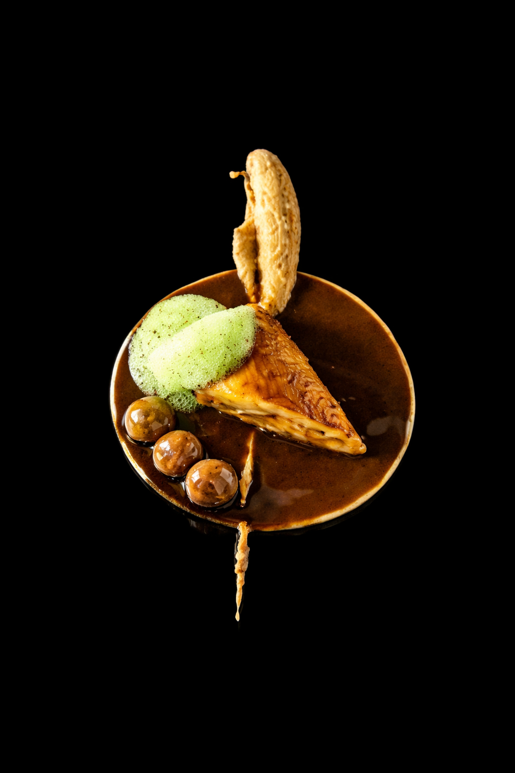 Un dessert avec une tranche de gâteau au caramel, de la glace verte, des billes en chocolat et un biscuit sec, servi dans un bol en chocolat sur un fond noir.