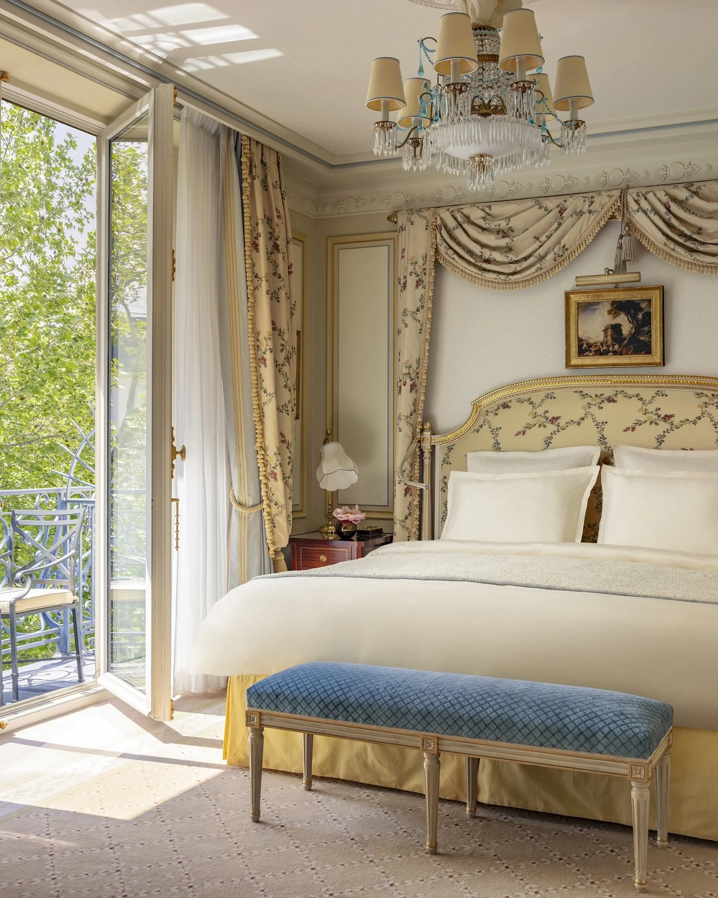 Chambre élégante avec un grand lit à tête rembourrée, rideaux fleuris, un petit banc au pied du lit, un lustre en cristal au plafond, et une porte-fenêtre donnant sur un balcon avec vue sur les arbres.