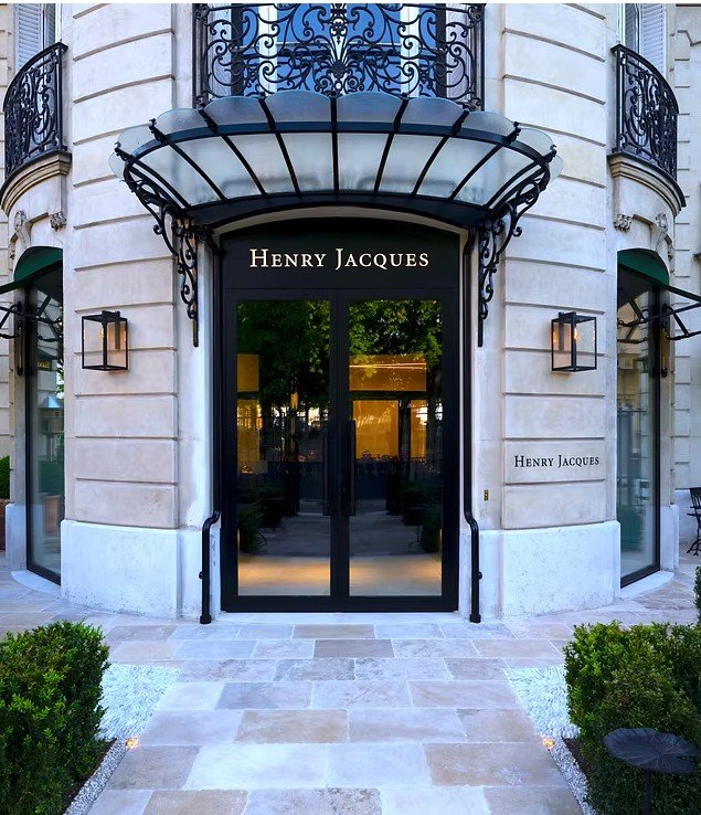 Entrée d'un bâtiment avec une porte en verre noir et le nom 'Henry Jacques' au-dessus. La façade n° 3, deux fenêtres avec volets verts, murs en pierre beige. Décorations en fer forgé au-dessus de la porte, deux lampes extérieures, petits buissons à gauche et à droite, pavage en pierre devant.