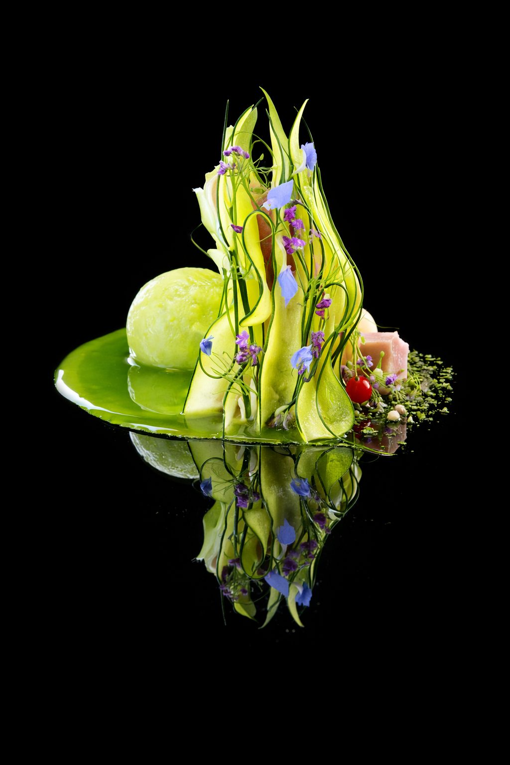 Une composition artistique de légumes et fleurs sur une surface noire, comprenant un melon vert, des courgettes, des fleurs violettes et bleues, et une base en forme de plaque