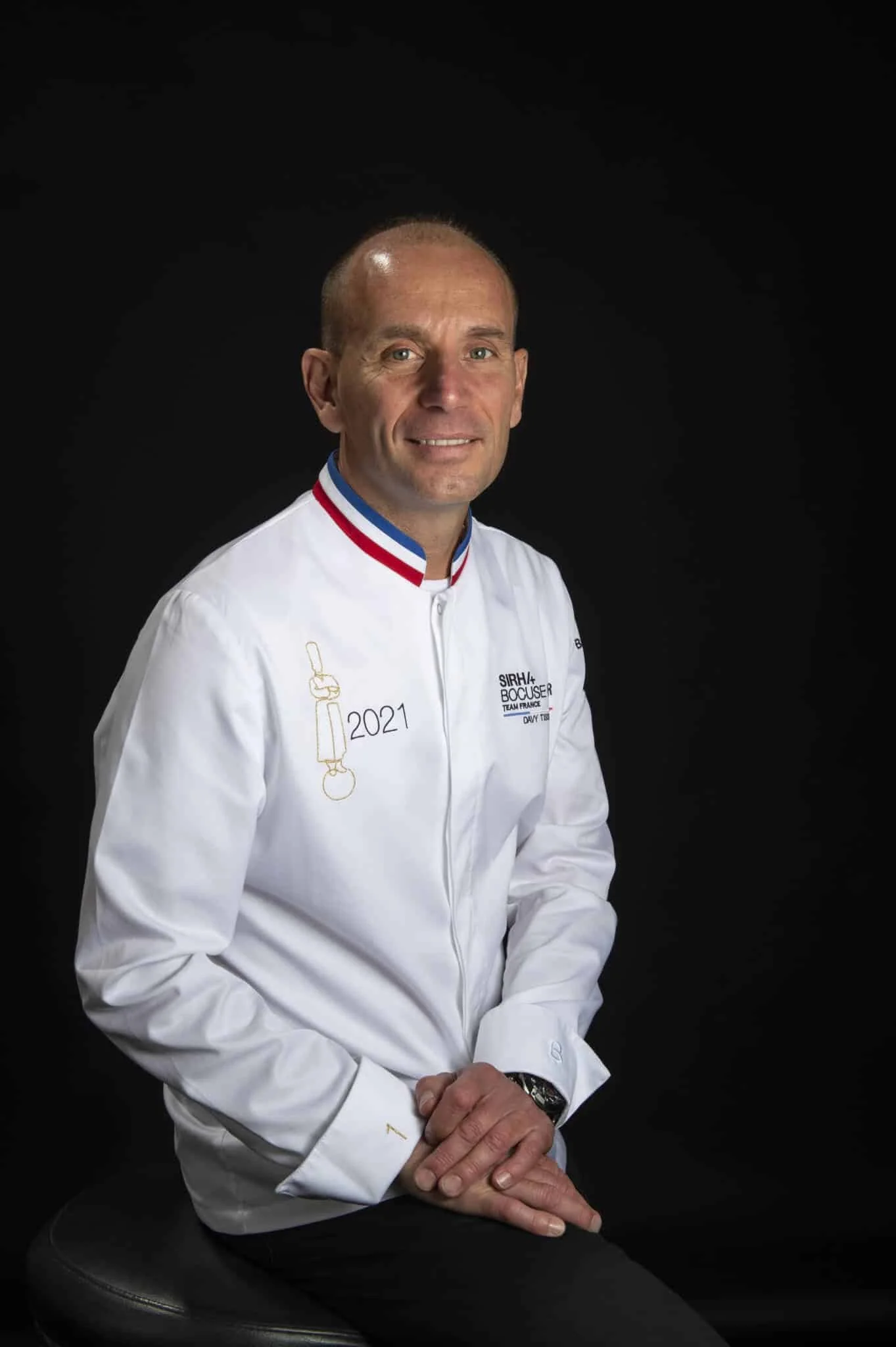 Homme en veste blanche avec un col tricolore, assis sur un tabouret noir contre un fond noir, portant une montre, souriant, avec un logo de chef cuisinier et une décoration '2021' sur la veste.