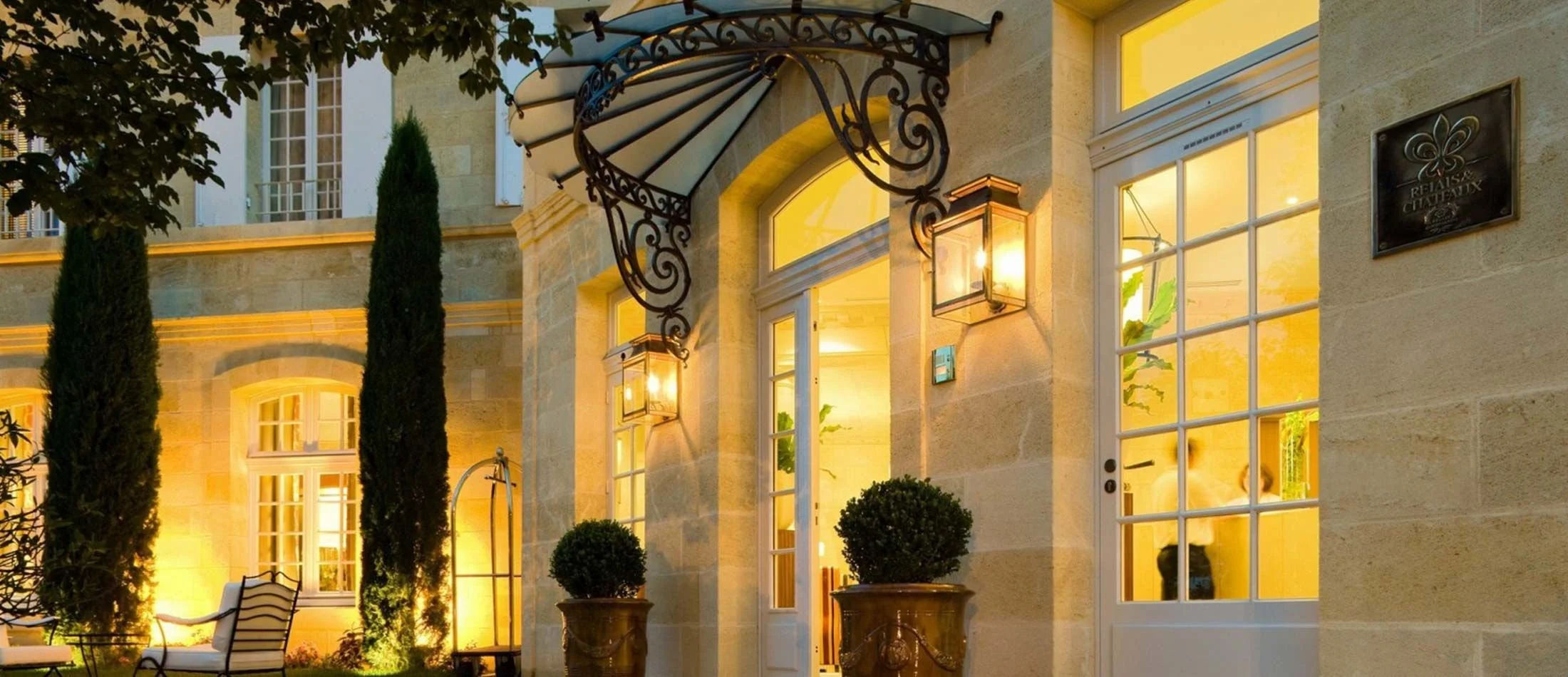 Facade d'une maison élégante en pierre, éclairée à la tombée de la nuit, avec des portes en verre et de la végétation ornementale, ambiance chaleureuse et invitante.