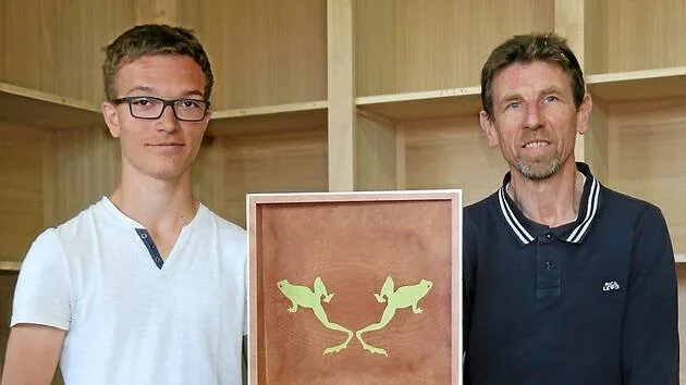 Deux hommes posent avec un tableau contenant deux figures de lézards verts. Un homme jeune porte un t-shirt blanc, un autre homme porte une chemise bleue, tous deux souriants.