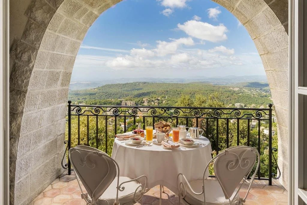 Petit déjeuner sur un balcon avec vue sur la nature, comprenant un tableau rond avec des verres de jus, assiettes, tasses, un cornet de croissants et une théière, entouré de deux chaises blanches.