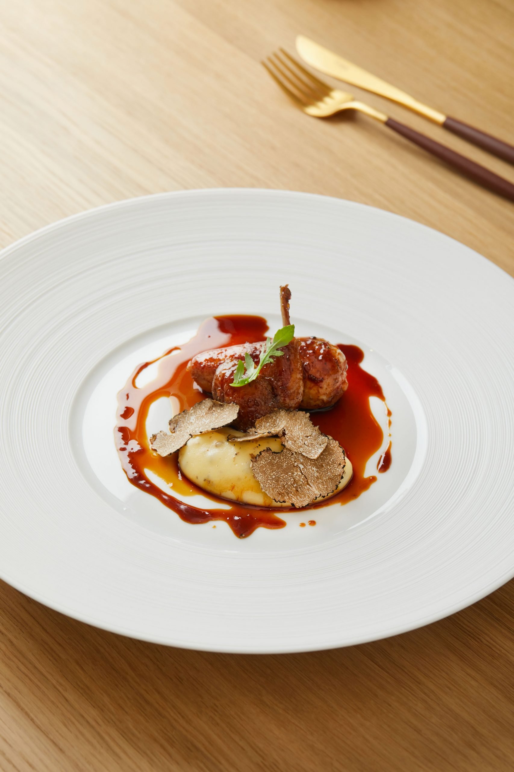 Assiette gastronomique avec un morceau de viande, sauce, truffes et purée, garnie d'une petite herbe, sur une table en bois avec des couverts en or et en bois à l'arrière-plan.