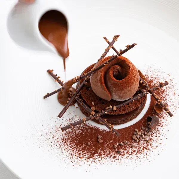 Une dessert à base de boule de glace ou mousse au chocolat, décorée avec des bâtons de chocolat, de la poudre de cacao, du cacao en poudre, et un filet de sauce chocolat being versé dessus.