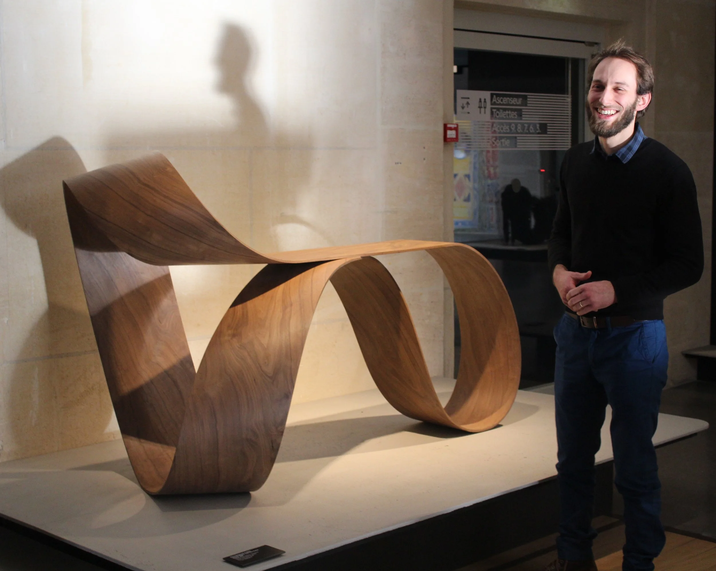Un homme souriant debout à côté d'une sculpture en bois courbée, exposée dans une galerie ou un musée.