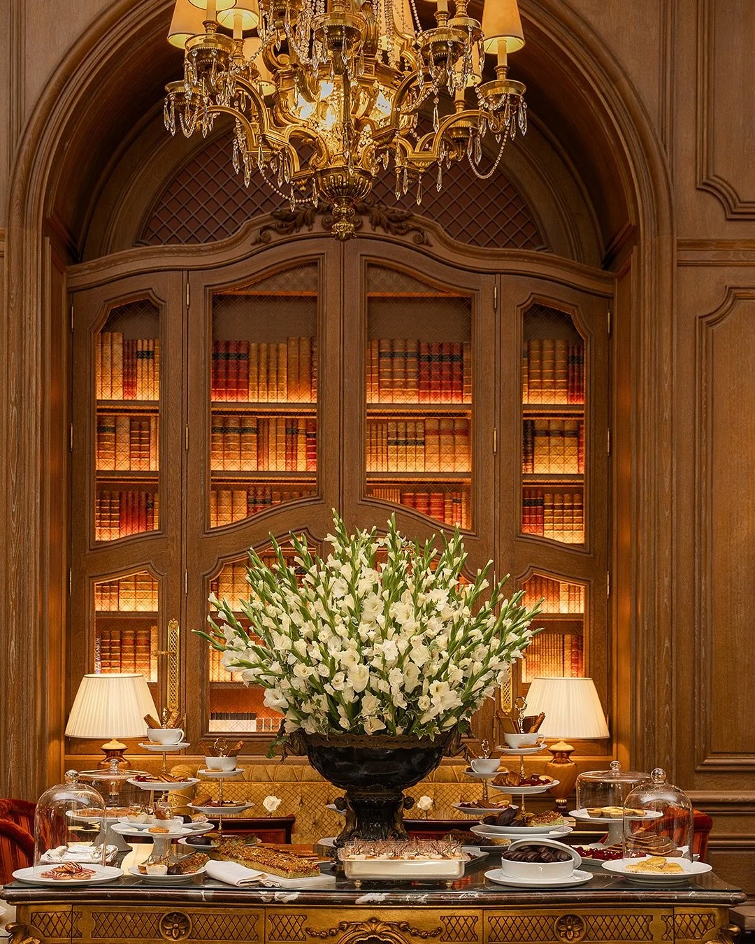 Une grande pièce élégante avec un bureau en bois richement décoré, une grande coupelle noire remplie de fleurs blanches, plusieurs lampes et une grande bibliothèque en arrière-plan.