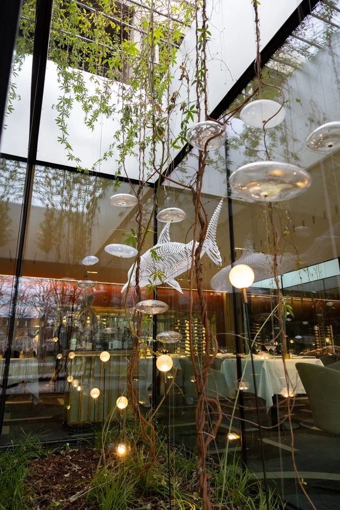 Vitrine de restaurant avec une décoration intérieure comprenant une sculpture de poisson en fil métallique suspendue et des lampes flottantes. Des plantes grimpantes pendent du plafond, créant une ambiance naturelle et moderne.