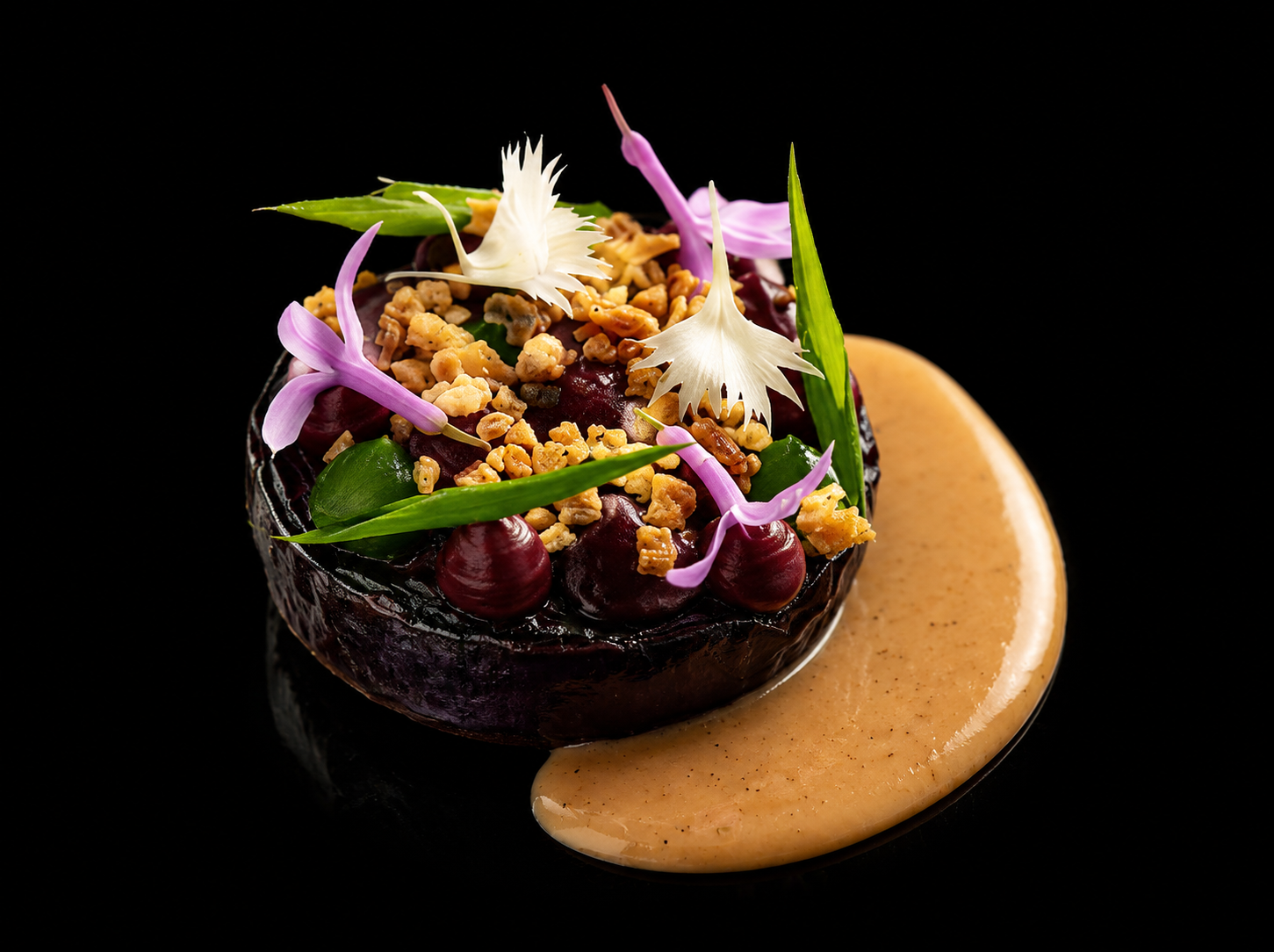 Plat de cuisine sophistiquée avec un morceau de légume (probablement une betterave) garnie de graines, fleurs comestibles, et feuilles, avec une sauce onctueuse beige à côté, sur fond noir.