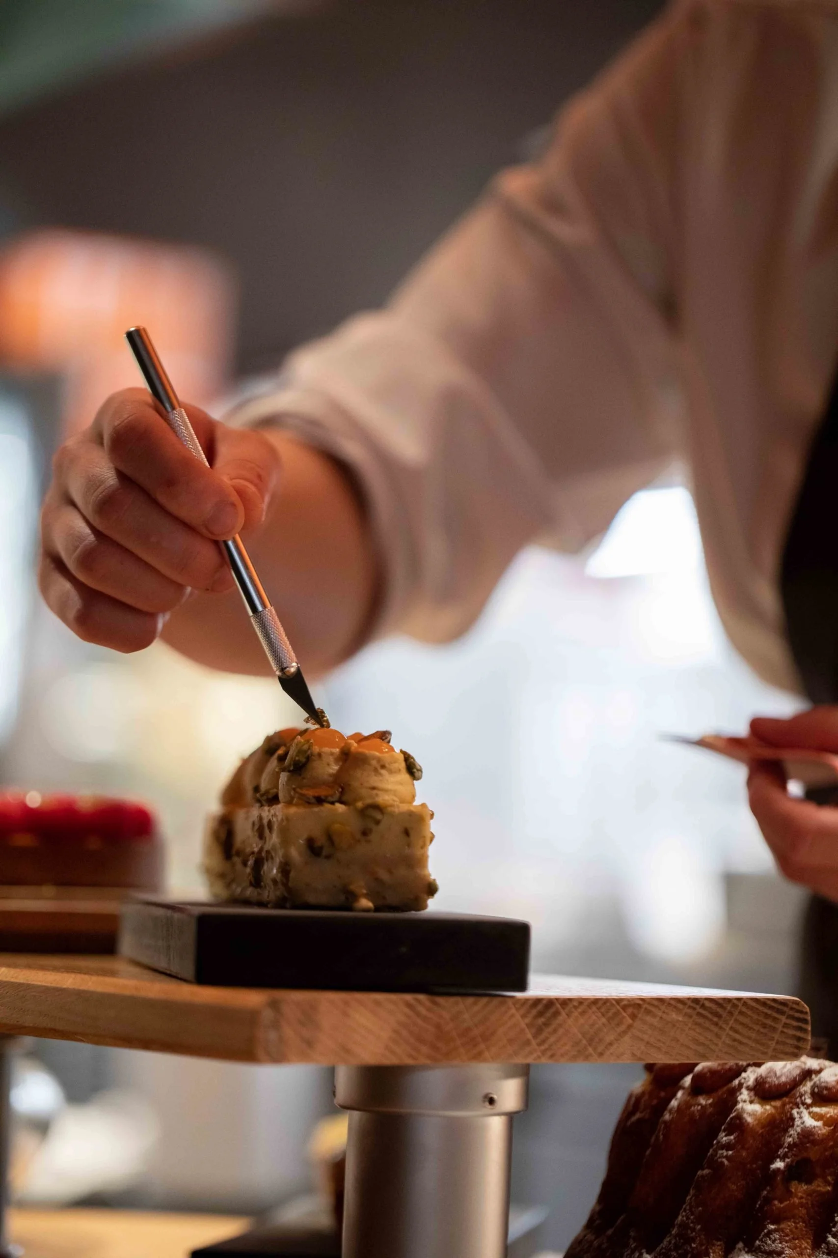 Une personne en train de décorer une pâtisserie avec un stylo de décoration, sur un plan de travail en bois. Il y a d'autres desserts visibles en arrière-plan.