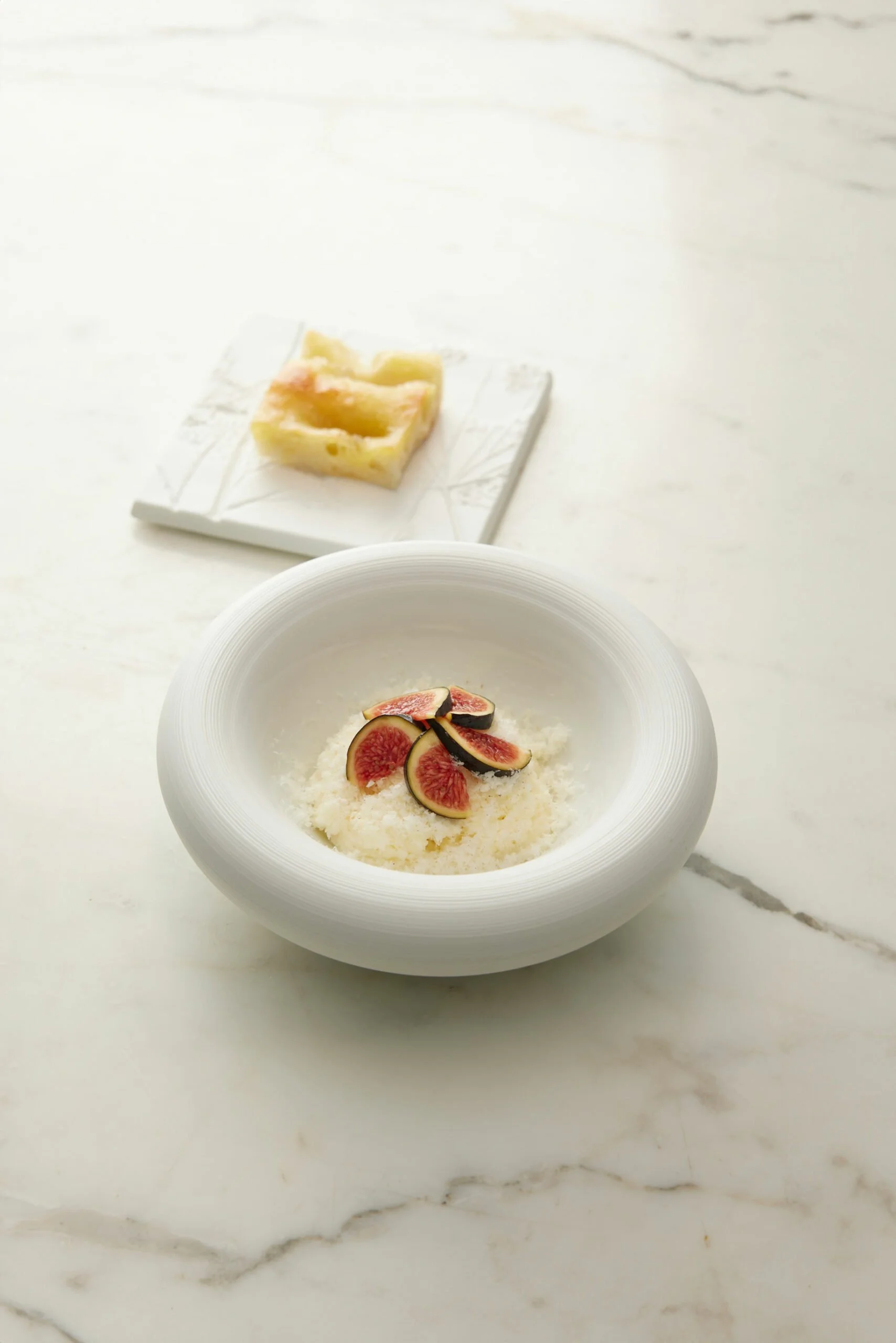 Une coupe blanche contenant des figues fraîches accompagnées de fromage râpé, avec un morceau de fromage sur une petite assiette en arrière-plan.