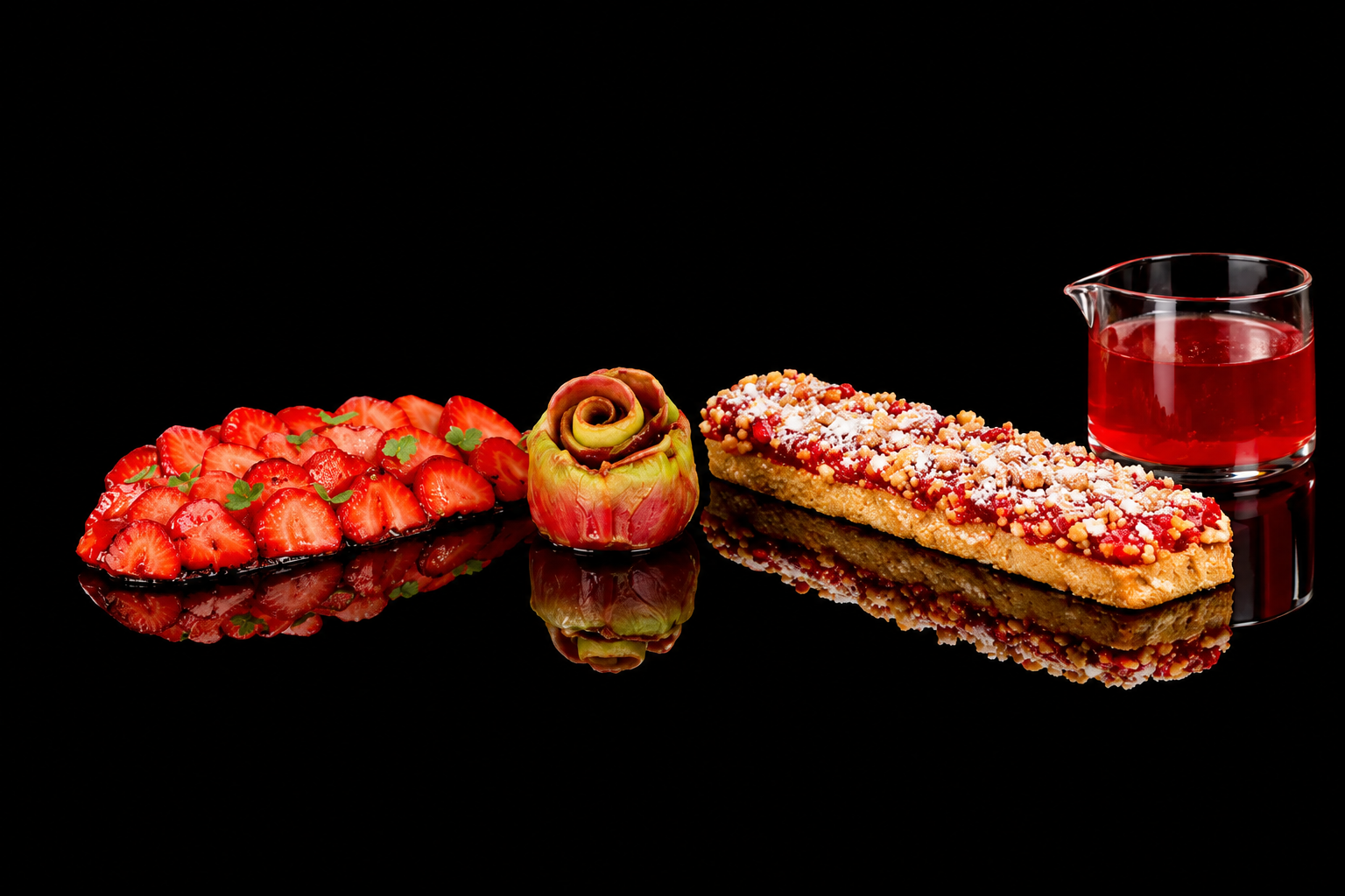 Groupe de desserts et de fruits, comprenant des fraises tranchées, une pomme transformée en décoration avec une torsade enroulée, une tarte ou gâteau aux fruits recouvert de sucre en poudre, et un petit pichet de sirop rouge, tous sur un fond noir réfléchissant.