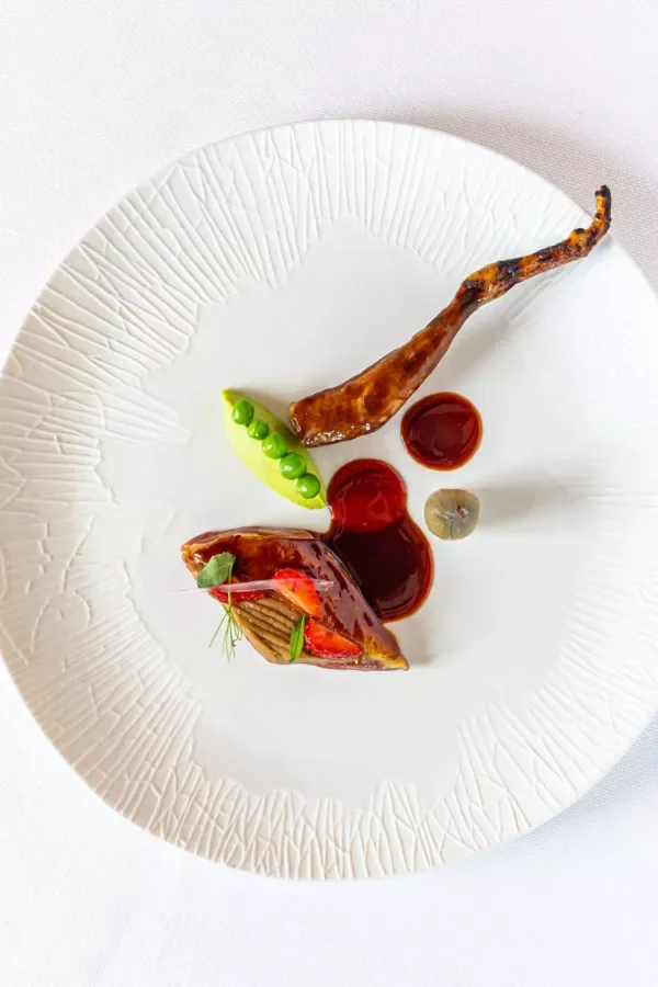 Assiette blanche avec un plat gastronomique composé de morceaux de viande, purée de petits pois, et deux sauces rouge foncé.